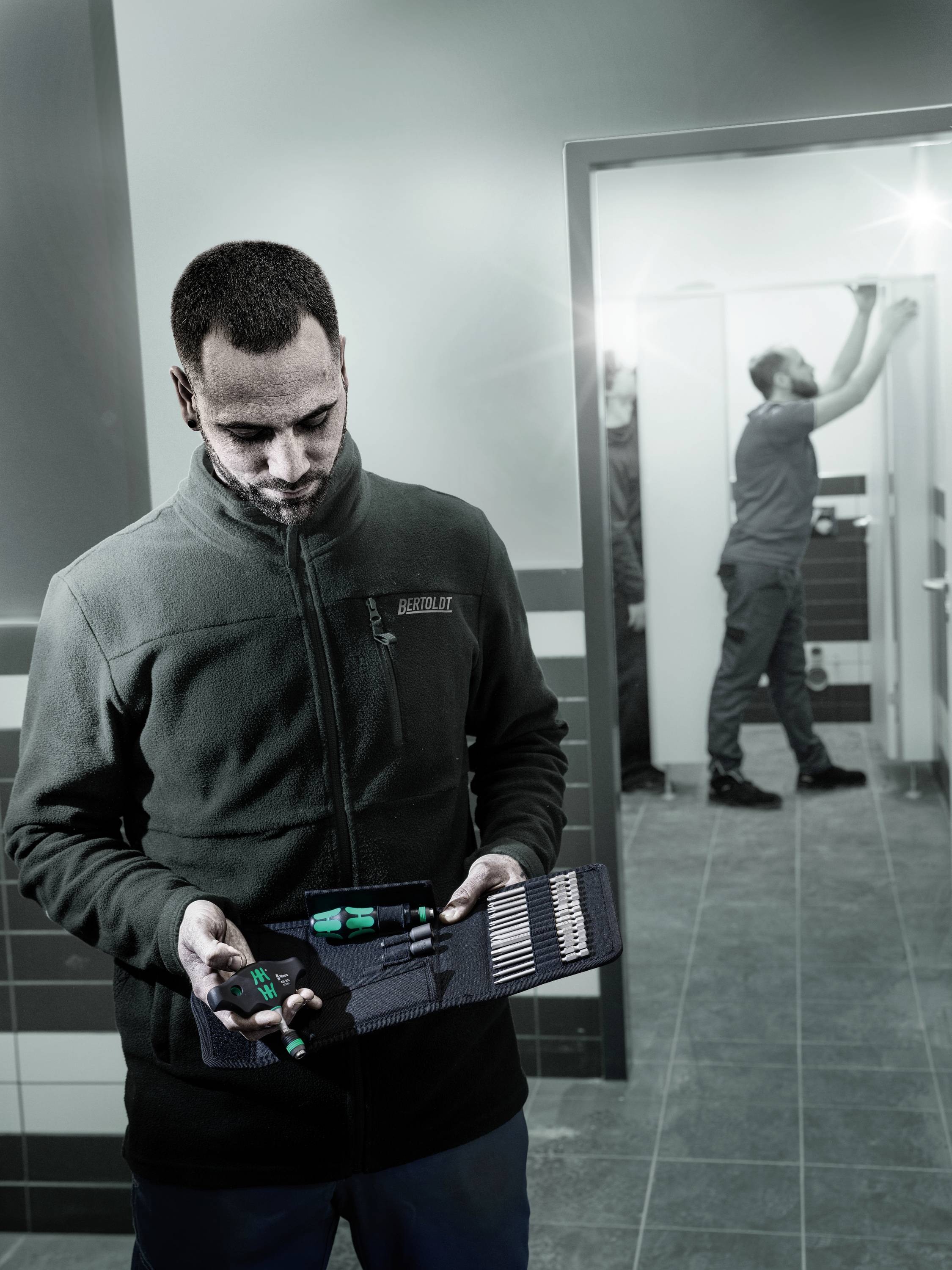 Ein Mann hält einen Werkzeugkoffer, während ein zweiter Mann im Hintergrund an der Decke arbeitet. Sie sind in einem hellen Raum.