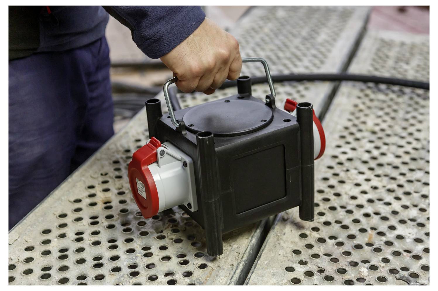 Hand, die eine tragbare elektrische Pumpe oder einen Generator auf einer perforierten Metalloberfläche im Freien hält.