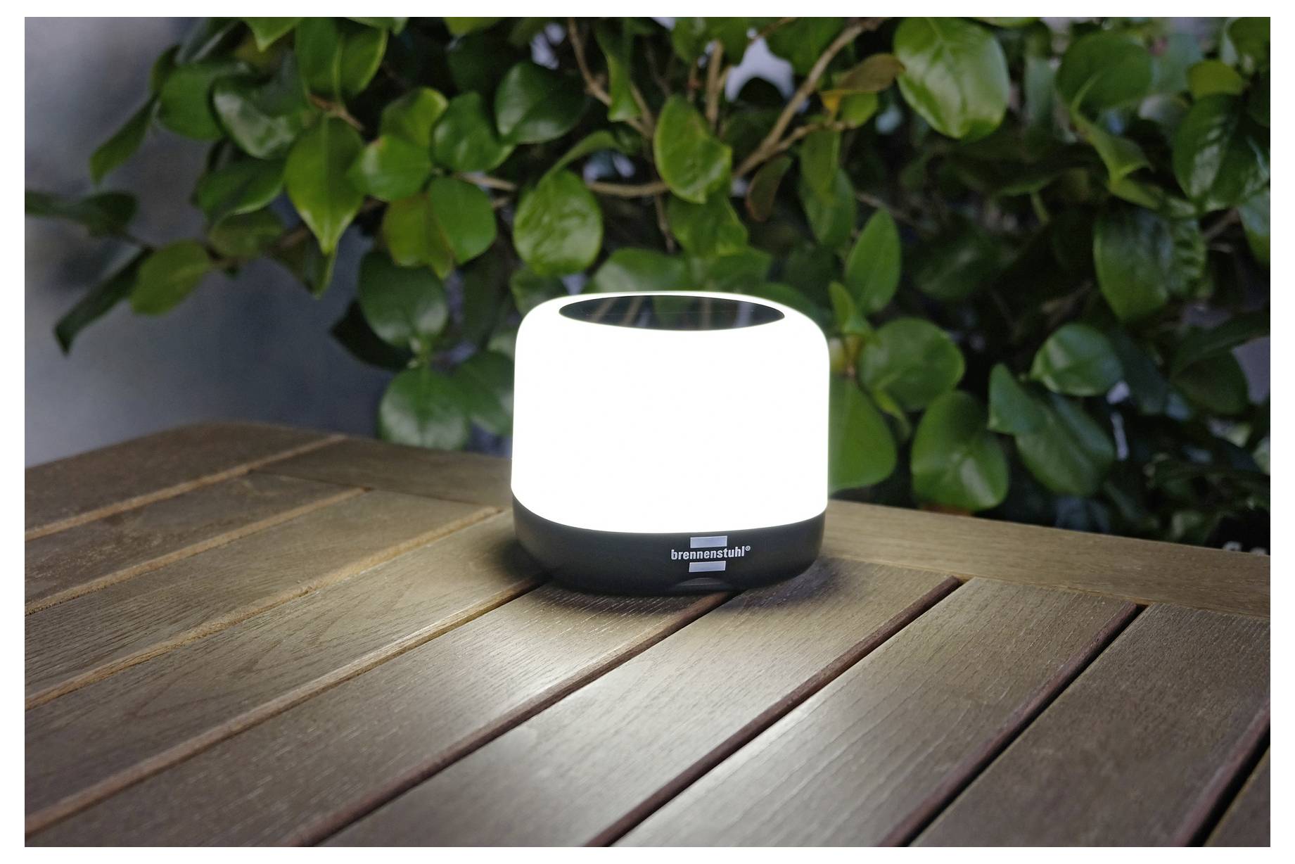 Kleine weiße zylindrische Lampe, die auf einem Holztisch leuchtet, mit grünem Laub im Hintergrund.
