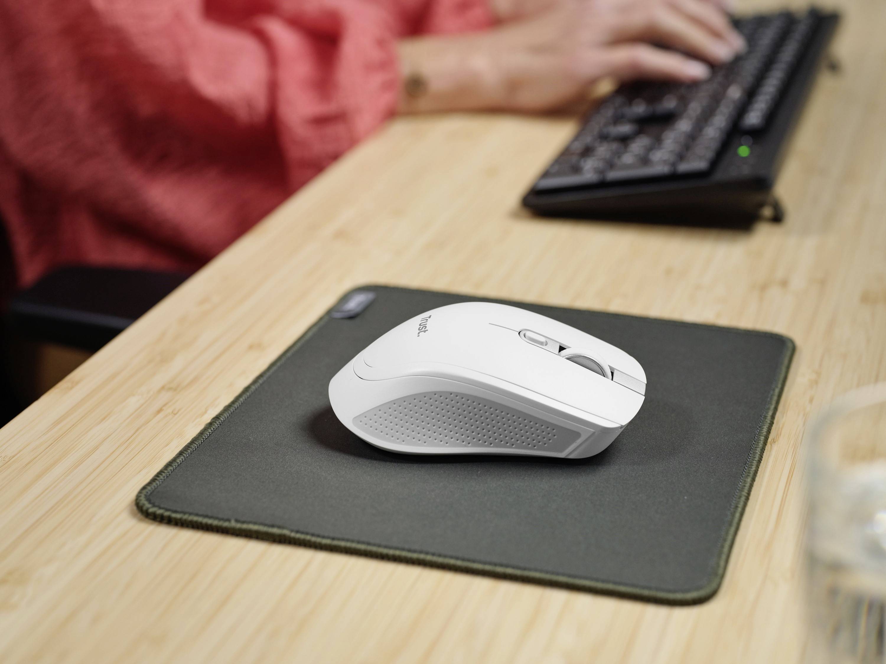 Eine weiße Computermaus auf einem grünen Mauspad, eine Person tippt auf einer Tastatur im Hintergrund.