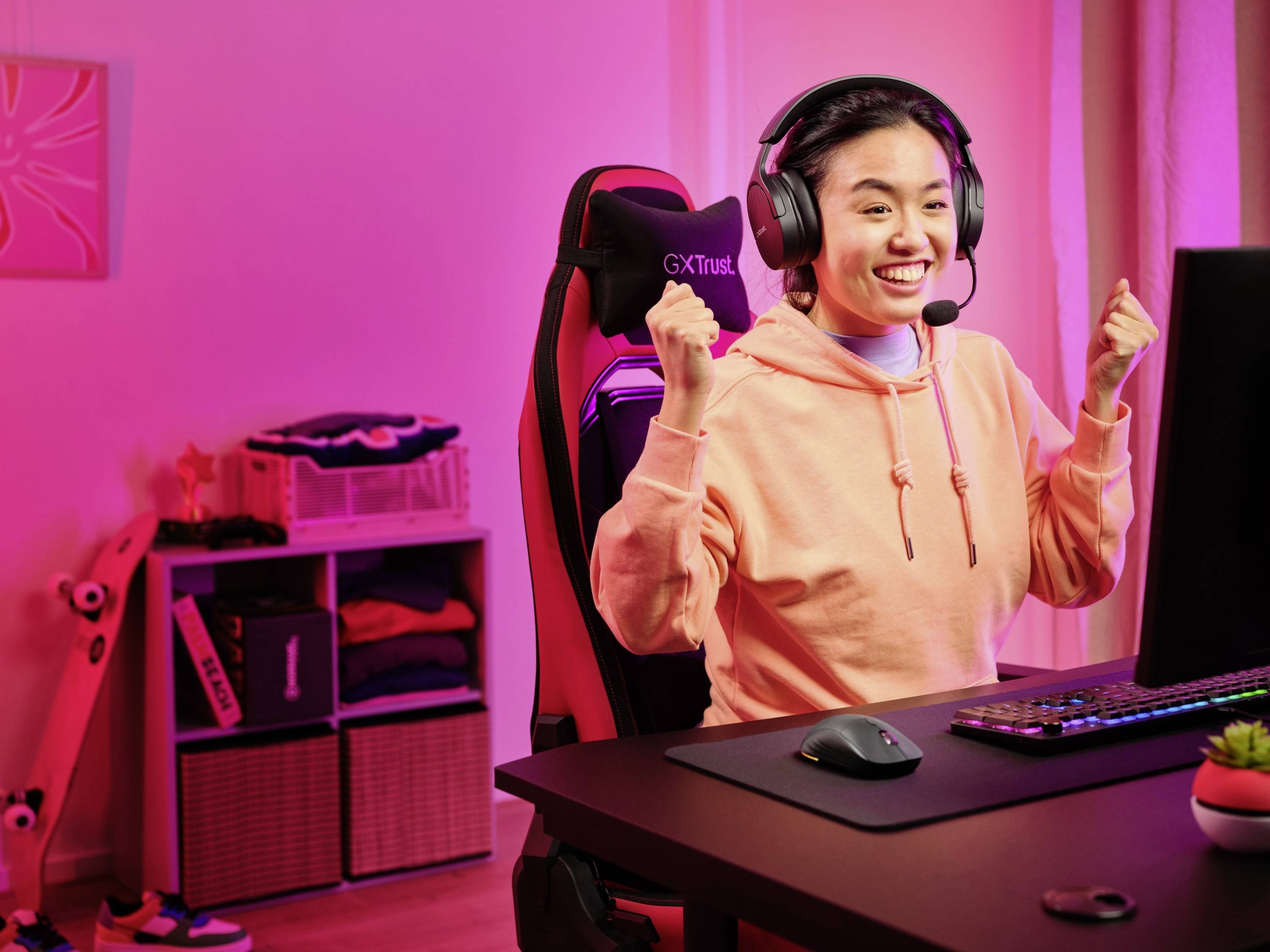 Eine Person sitzt vor einem Computer in einem pink beleuchteten Raum, trägt ein Headset und freut sich mit erhobenen Fäusten.