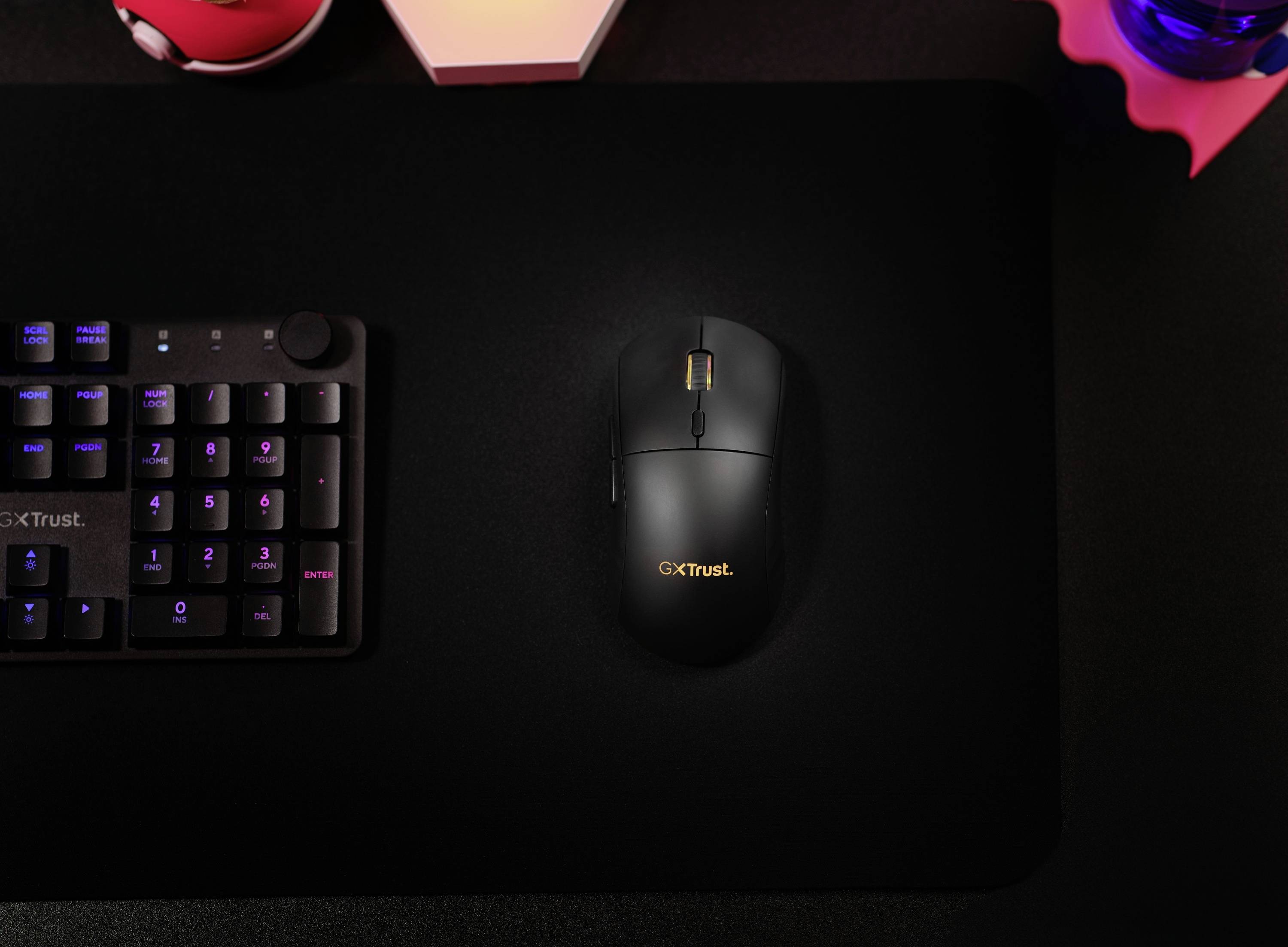Eine schwarze Computermaus mit der Aufschrift 'GxTrust' auf einem dunklen Mauspad neben einer Tastatur auf einem Schreibtisch.