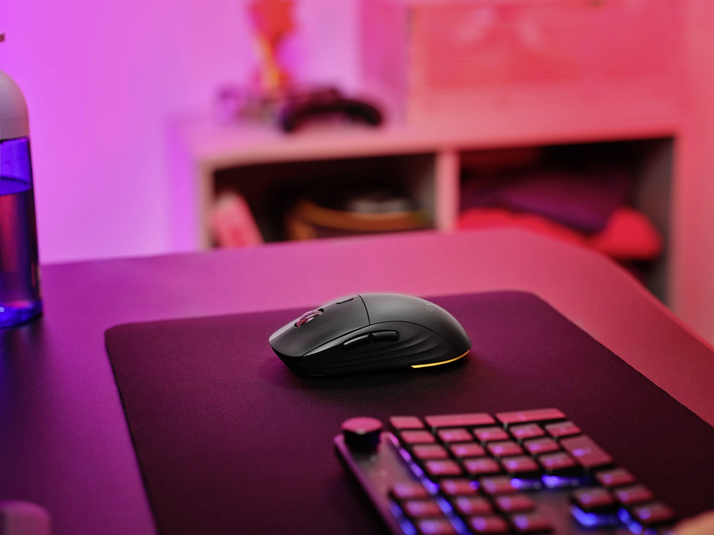 Eine kabellose Maus auf einem schwarzen Mauspad, im Hintergrund ein Regal mit unscharfen Objekten. Raum in rosa Licht getaucht.