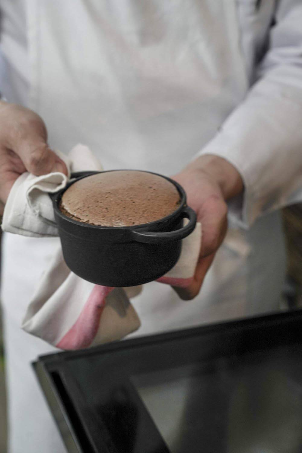Eine Person in weißer Kleidung hält einen warmen, frisch gebackenen Schokoladenkuchen in einer schwarzen Auflaufform mit einem Tuch.