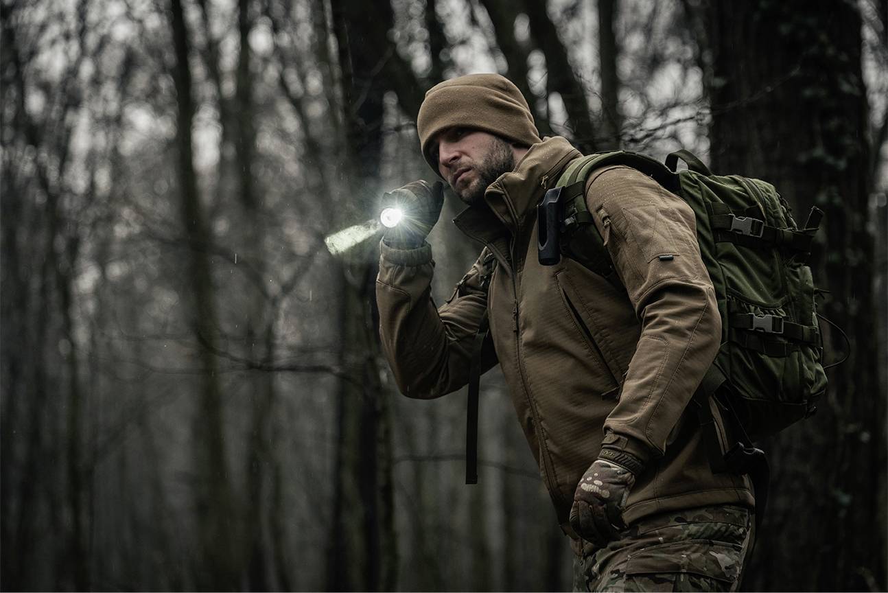 Eine Person in Outdoor-Kleidung und Mütze geht durch den Wald und hält eine Taschenlampe in der Hand. Es ist dunkel und nebelig.