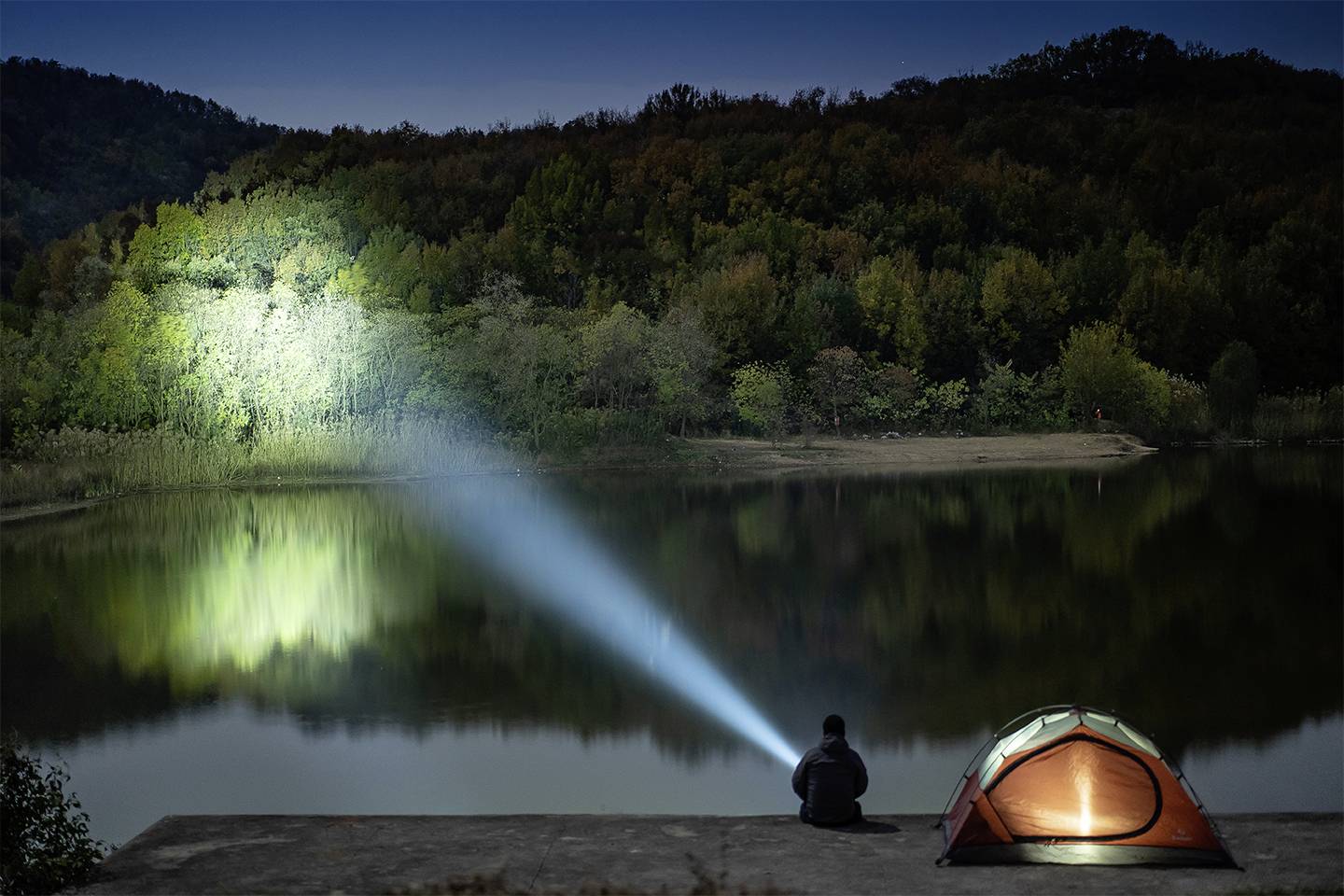 Ein Person sitzt nachts am See neben einem beleuchteten Zelt und richtet eine Taschenlampe auf die Bäume, die im Wasser gespiegelt werden.