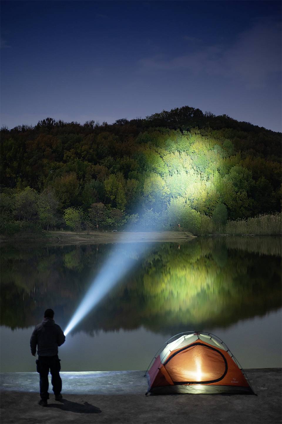 Eine Person steht mit einer Taschenlampe am Ufer eines Sees, beleuchtet ein Waldstück in der Nacht. Ein Zelt mit Licht steht daneben.