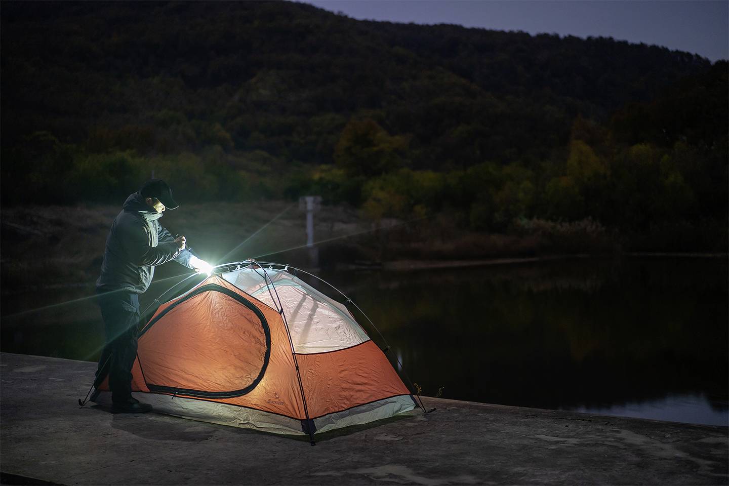 Eine Person baut nachts mit einer Taschenlampe ein orangefarbenes Zelt am Ufer eines ruhigen Sees vor bewaldeten Hügeln auf.