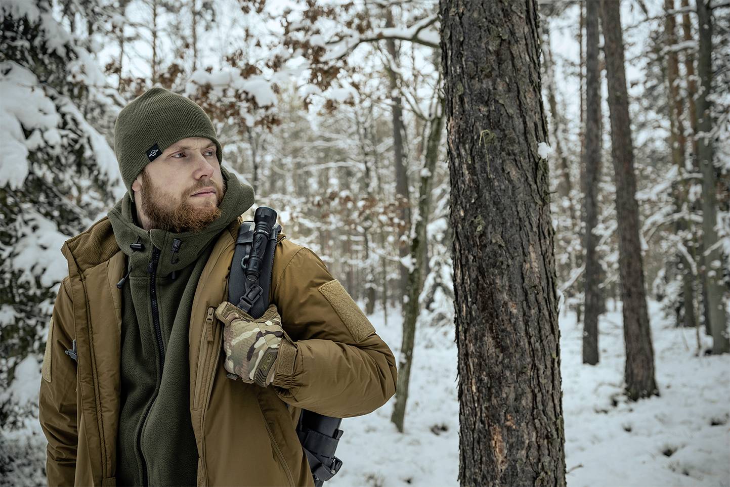 Ein Mann im Winter-Outfit, umgeben von verschneitem Wald, schaut zur Seite. Er hält einen schwarzen Rucksack und trägt Handschuhe.