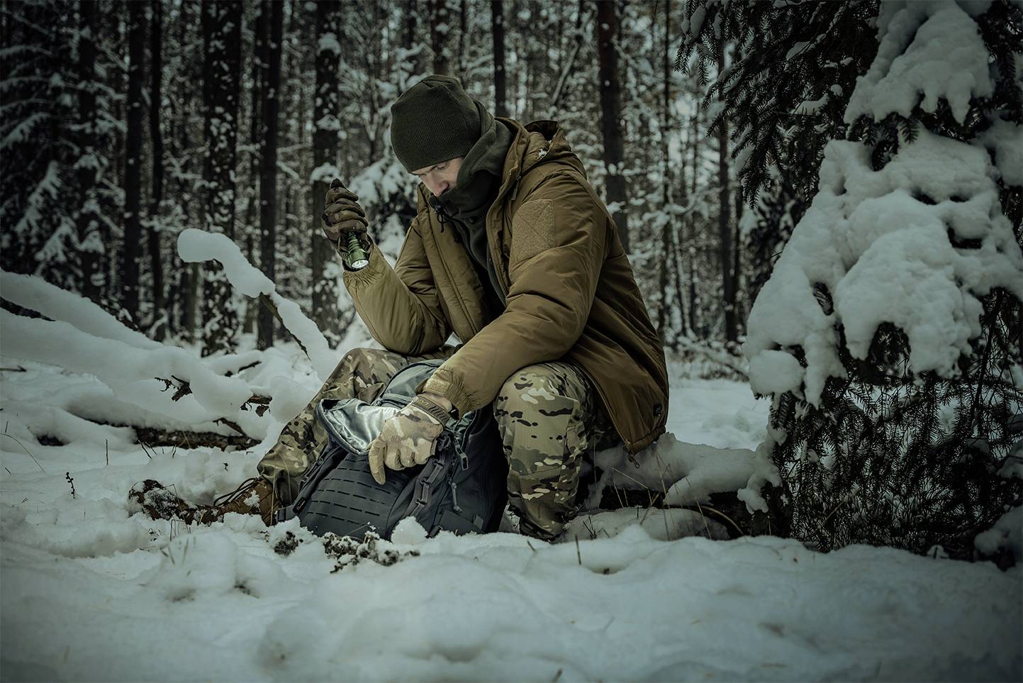 Eine Person sitzt in einem verschneiten Wald, trägt warme Kleidung, öffnet einen Rucksack. Umgebung ist voller Schnee und Nadelbäume.