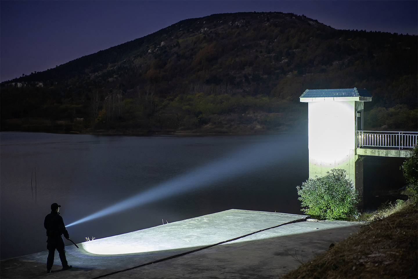 Ein Mann leuchtet nachts mit einer starken Taschenlampe auf einen Steg am See. Im Hintergrund ist ein bewaldeter Hügel zu sehen.