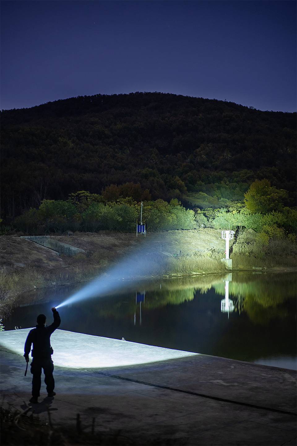 Eine Person leuchtet nachts mit einer Taschenlampe auf einen See, der von bewaldeten Hügeln umgeben ist. Reflektiertes Licht im Wasser.