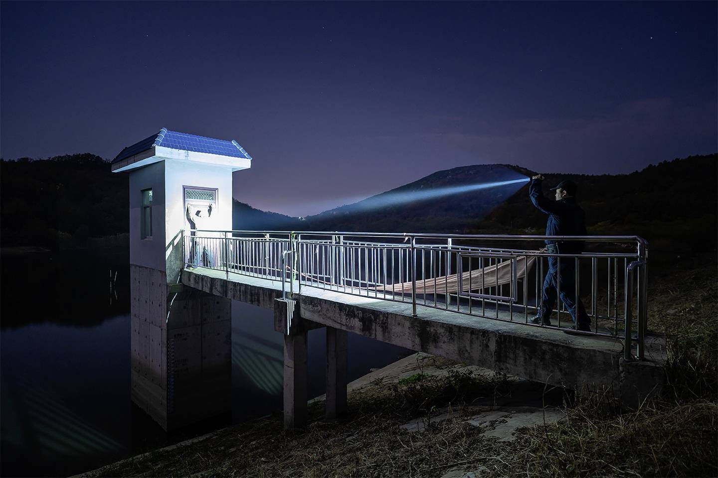 Eine Person beleuchtet nachts mit einer Taschenlampe die Seite eines Gebäudes auf einer Brücke über einem ruhigen Gewässer.
