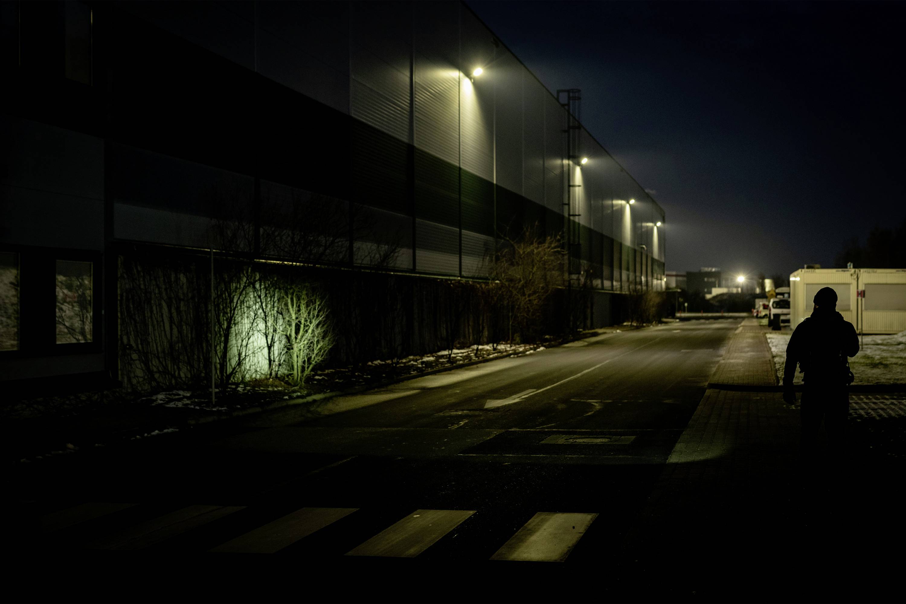 Eine dunkle Straße bei Nacht mit beleuchteten Gebäuden auf der linken Seite. Eine Person geht auf dem Bürgersteig entlang.