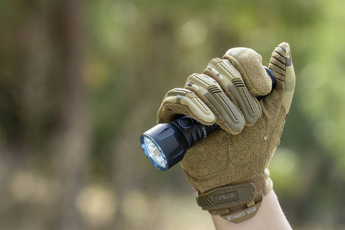 Eine Hand in einem braunen Handschuh hält eine eingeschaltete Taschenlampe im Freien. Der Hintergrund ist unscharf.