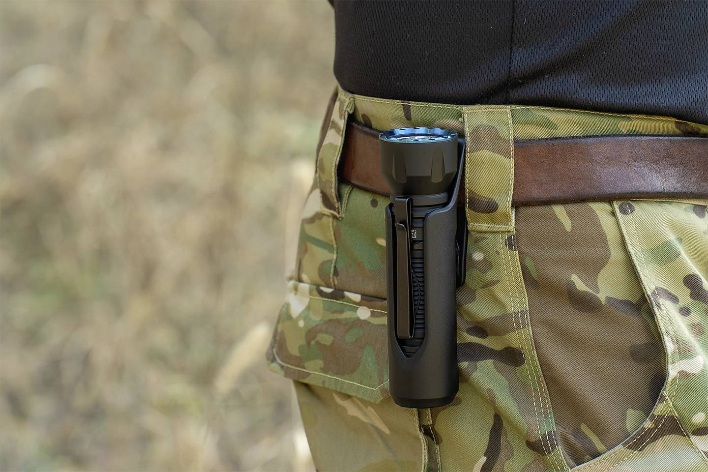 Person trägt eine Camouflage-Hose mit einer Taschenlampe im Holster an der Hüfte. Outdoor-Setting im Hintergrund.