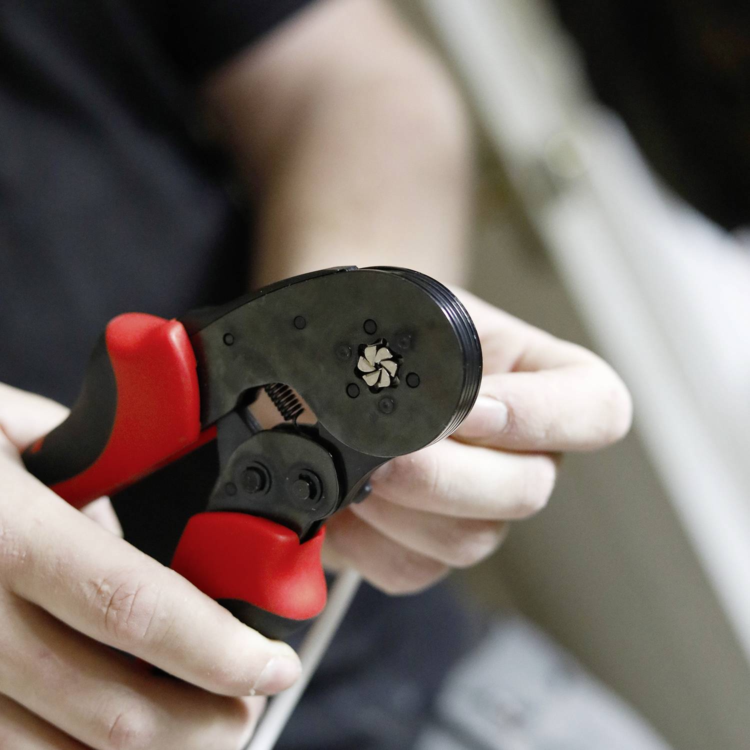 Eine Person bedient eine rote und schwarze Crimpzange, um ein Kabel zu bearbeiten. Die Szene zeigt eine Nahaufnahme der Hände bei der Arbeit.