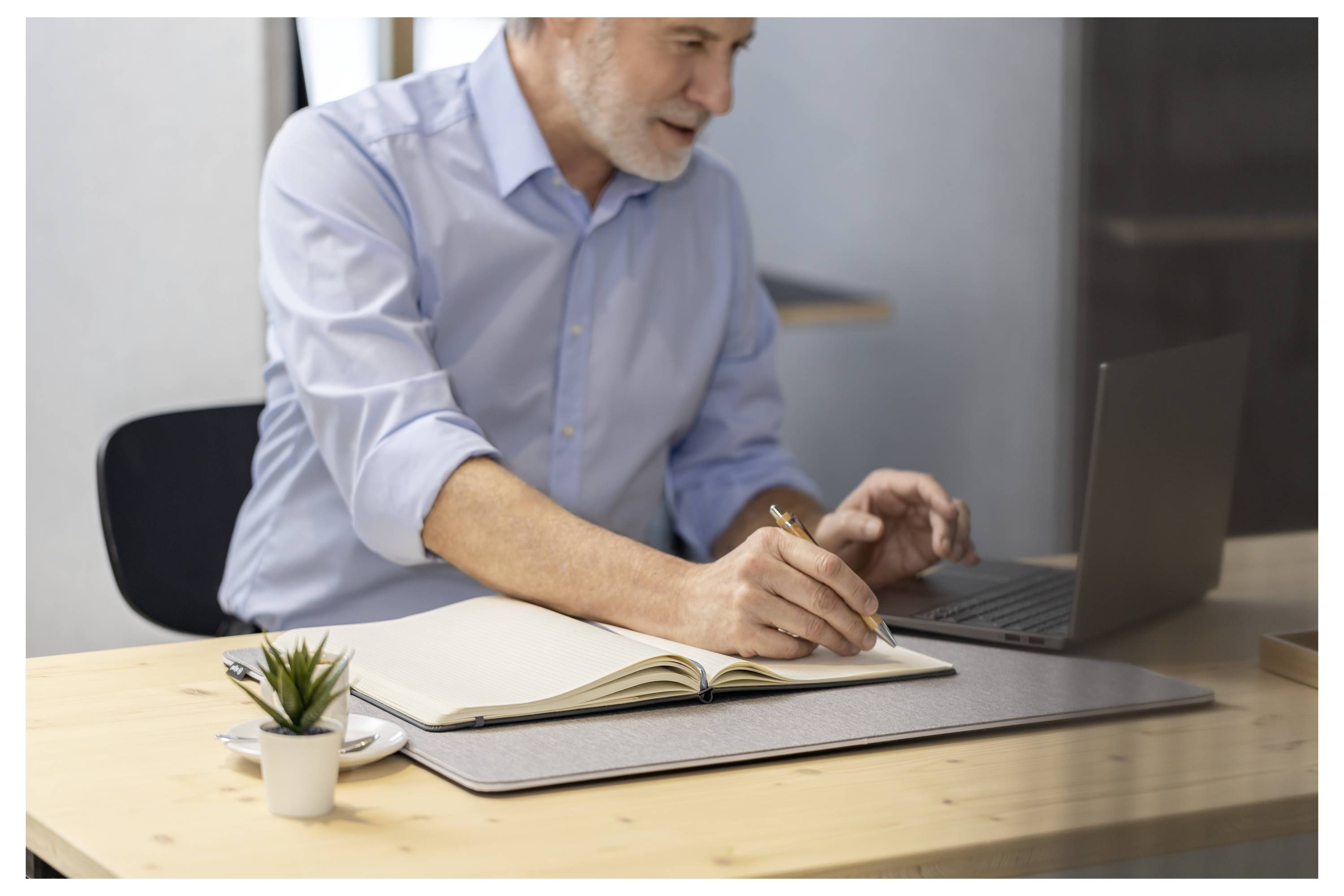 Ein älterer Mann in einem blauen Hemd schreibt in ein offenes Notizbuch, während er an einem Schreibtisch mit einem kleinen Laptop, einer Pflanze und einer Kaffeetasse arbeitet.