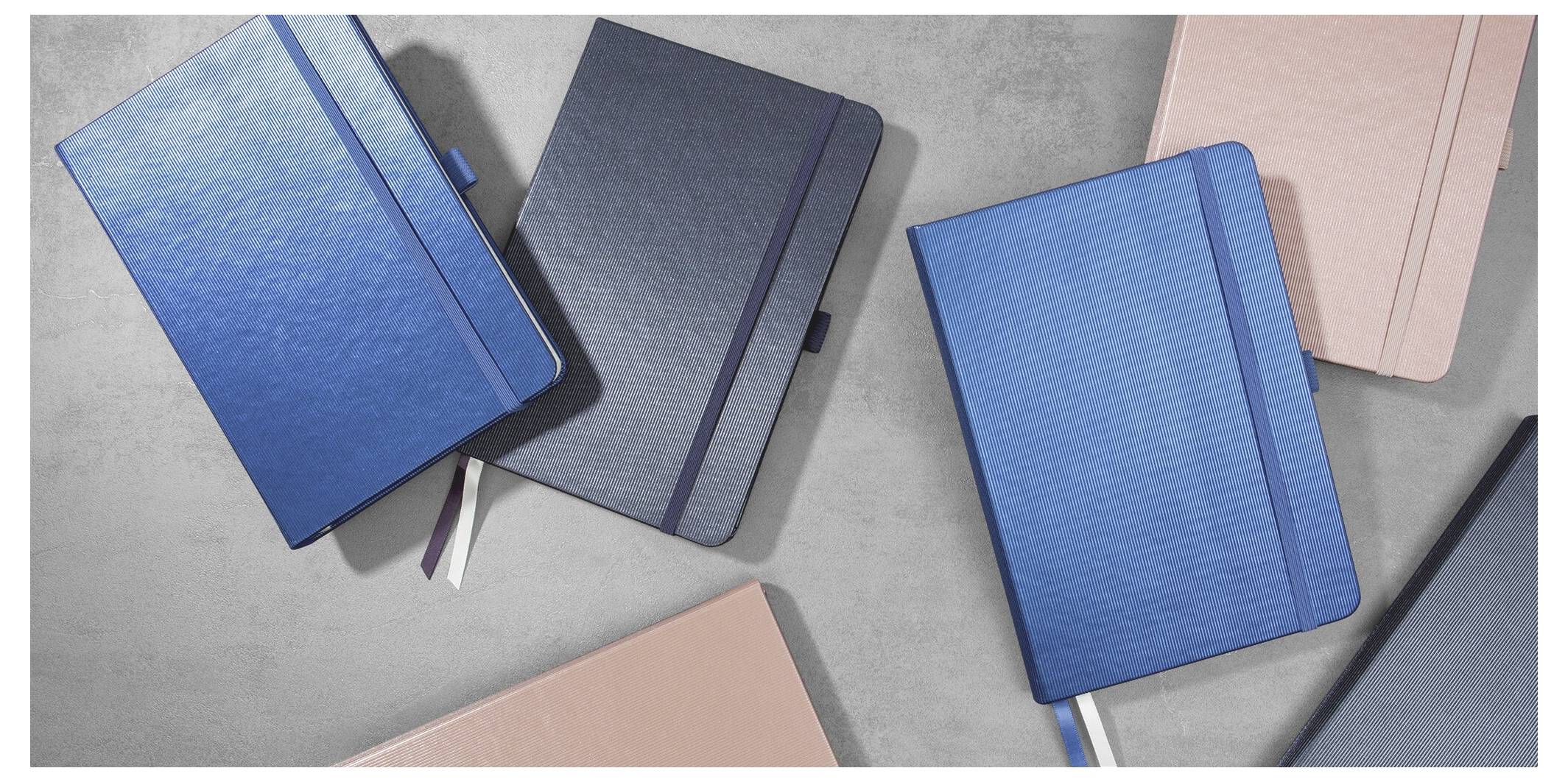 Vier geschlossene gebundene Notizbücher in Blau, Grau und Rosa diagonal auf einer hellgrauen Oberfläche angeordnet.