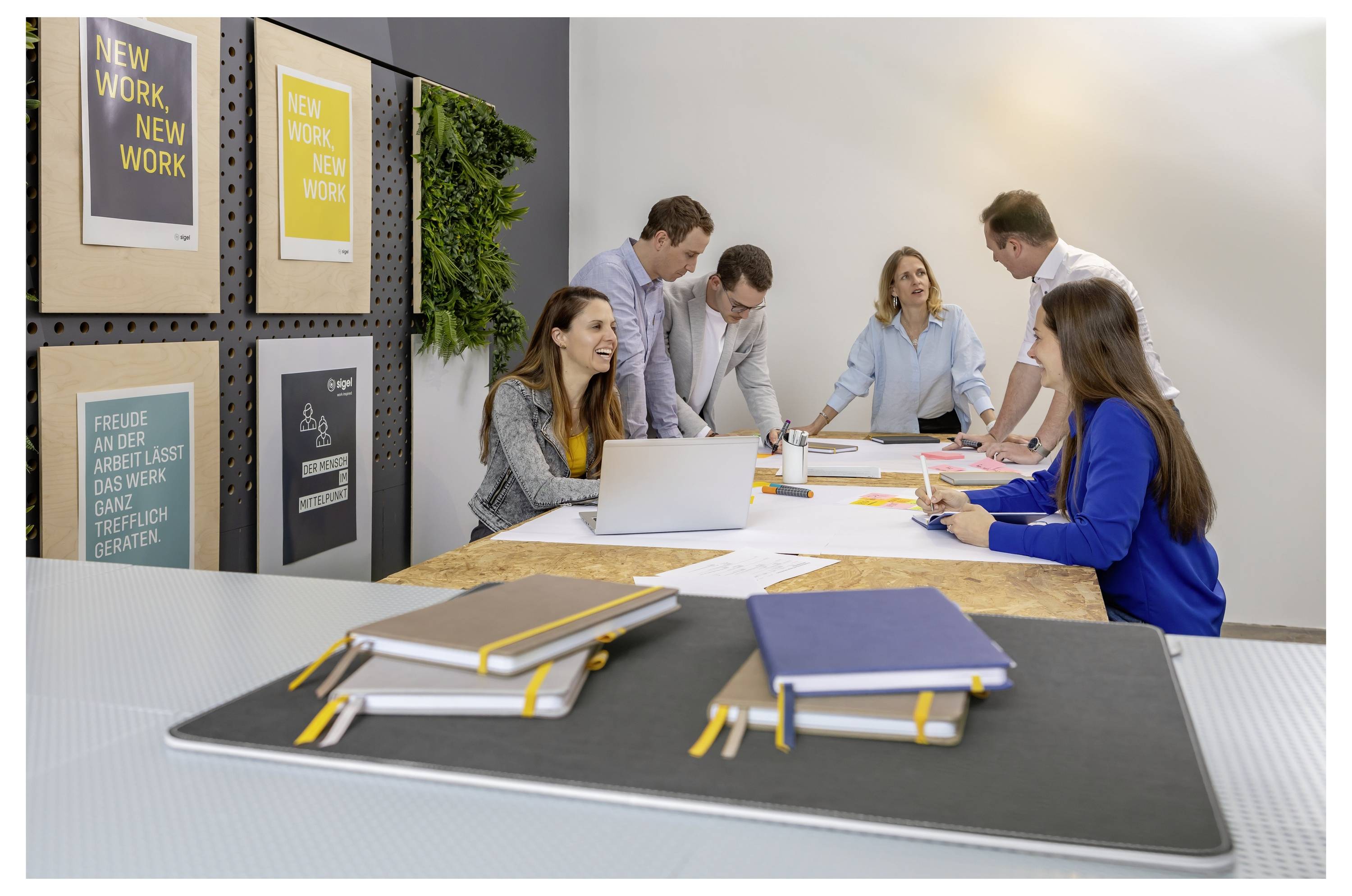 Geschäftsteam, das an einem Tisch in einem modernen Büro zusammenarbeitet, mit Notizbüchern im Vordergrund und motivierenden Wandpostern dahinter.