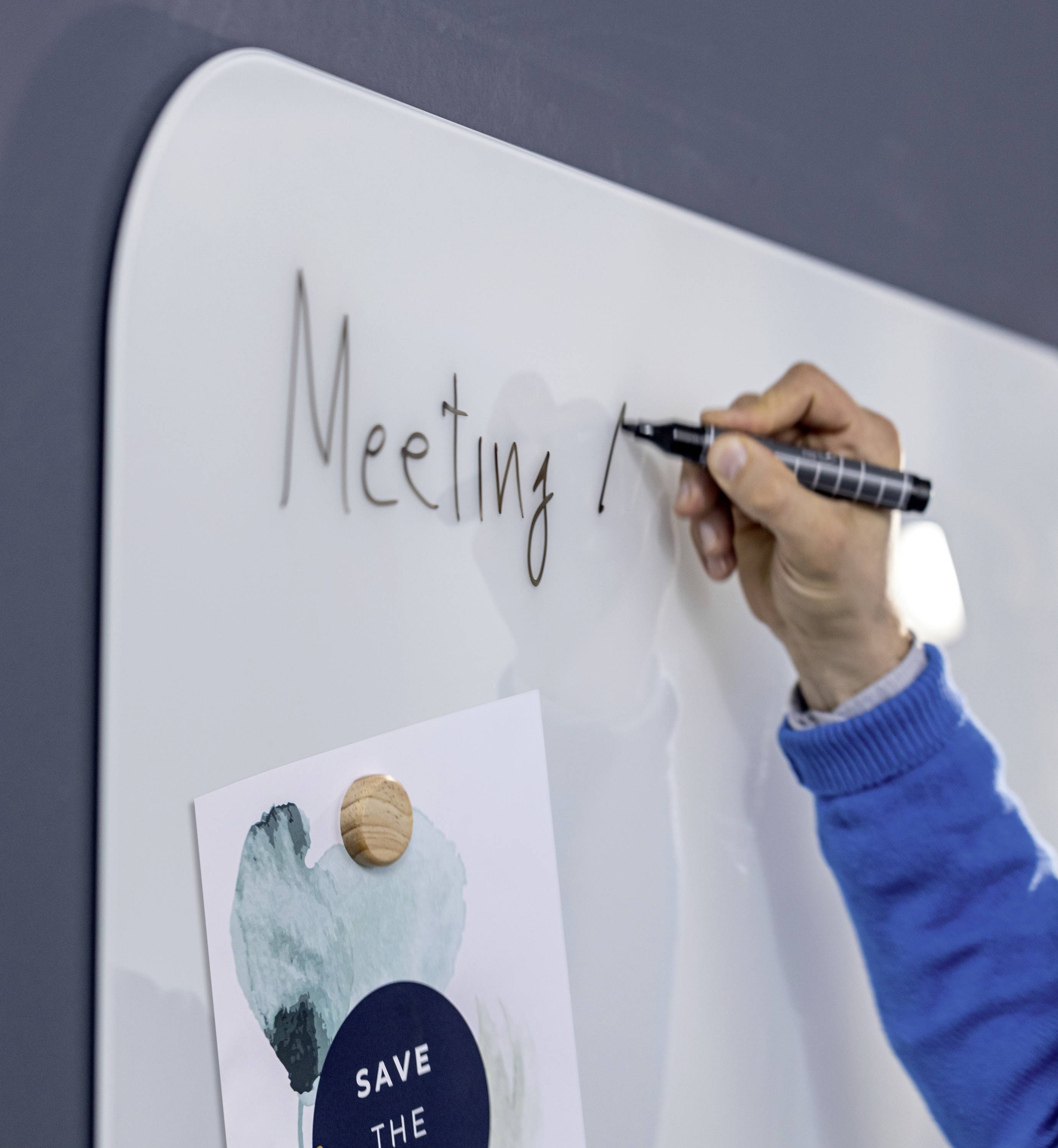 Eine Person schreibt 'Meeting!' auf ein Whiteboard. Darunter ist ein Blatt Papier mit der Aufschrift 'SAVE THE' und grünem Muster befestigt.