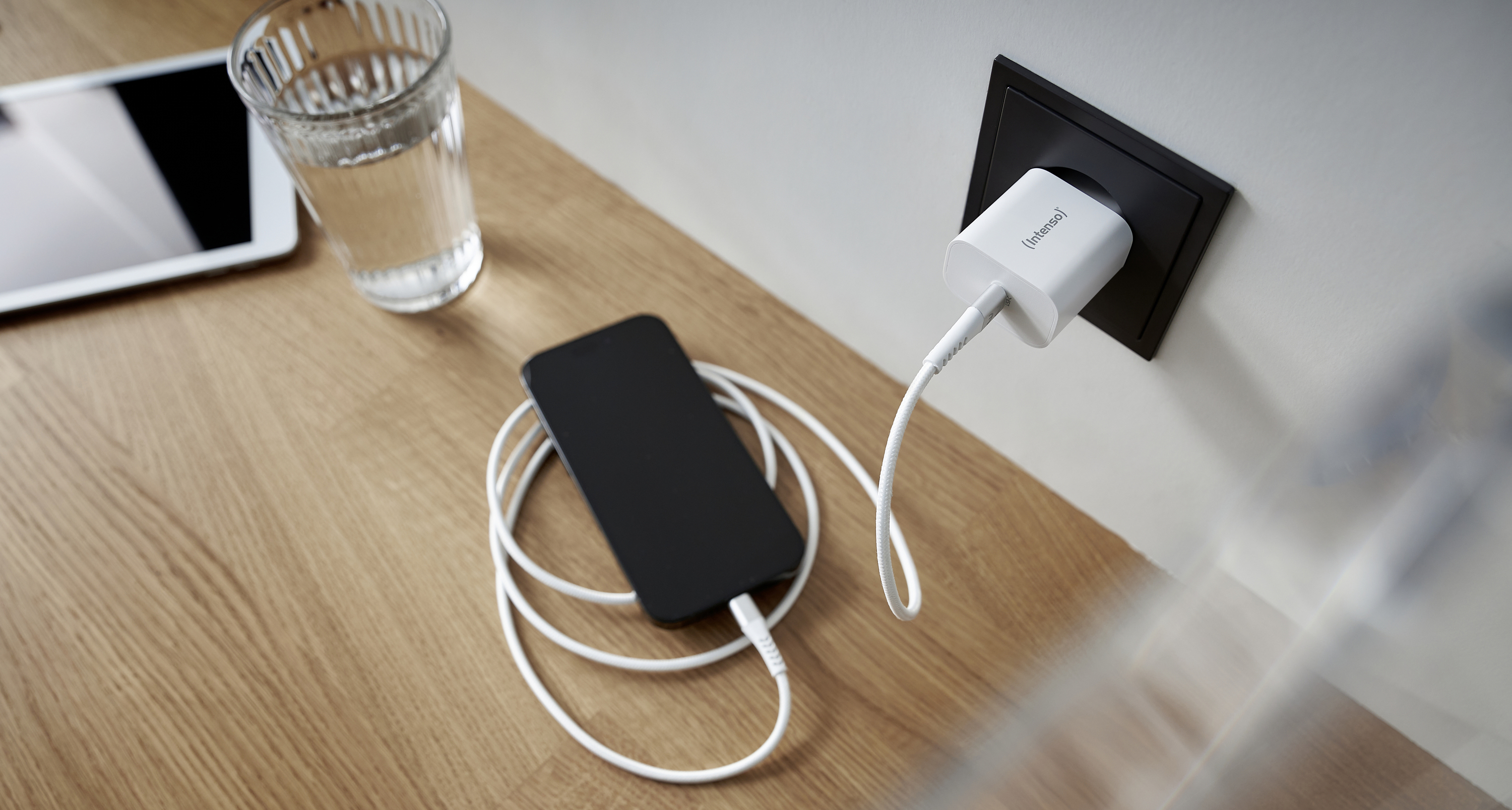 Ein Smartphone liegt auf einem Holztisch und wird über ein weißes Kabel an einer Steckdose geladen. Daneben ein Glas Wasser und ein Tablet.
