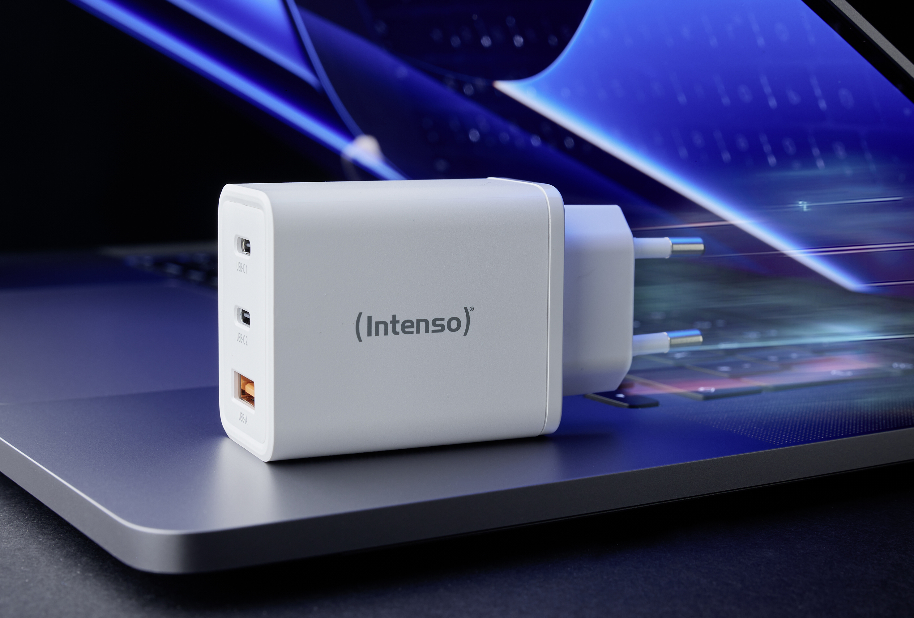Ein weißes Ladegerät mit der Aufschrift 'Intenso' liegt auf einem Laptop. Es verfügt über zwei USB-C- und einen USB-A-Anschluss.