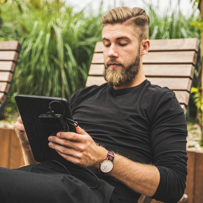 Ein Mann mit Bart liegt entspannt auf einer Sonnenliege und liest auf einem Tablet. Im Hintergrund sind Bäume und Pflanzen.