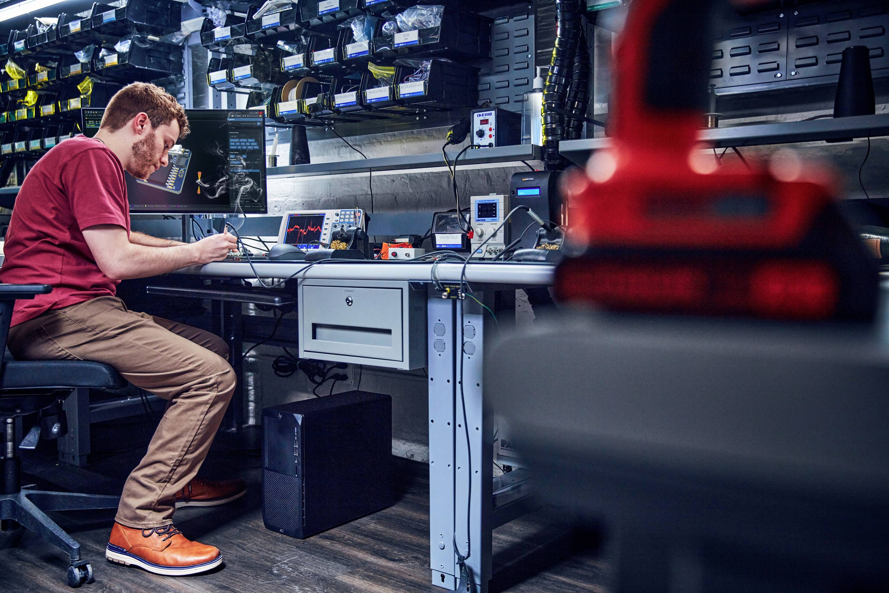 Ein Mann in einem Labor sitzt an einem Tisch und arbeitet an elektronischen Geräten. Der Raum ist mit Regalen voller Ausrüstung ausgestattet.