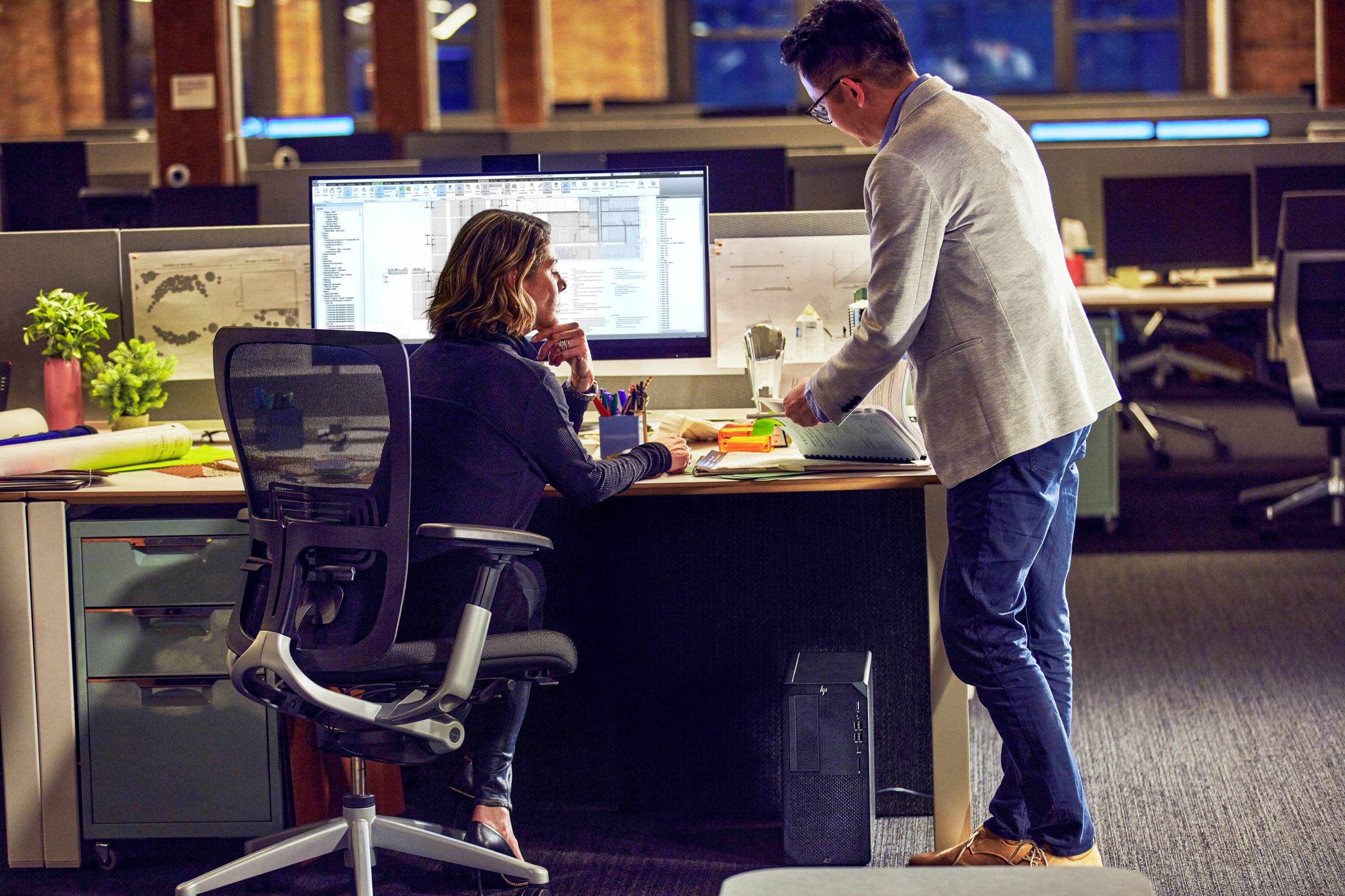 Zwei Personen arbeiten an einem Schreibtisch in einem modernen Büro, vertieft in ein gemeinsames Projekt auf einem großen Monitor.
