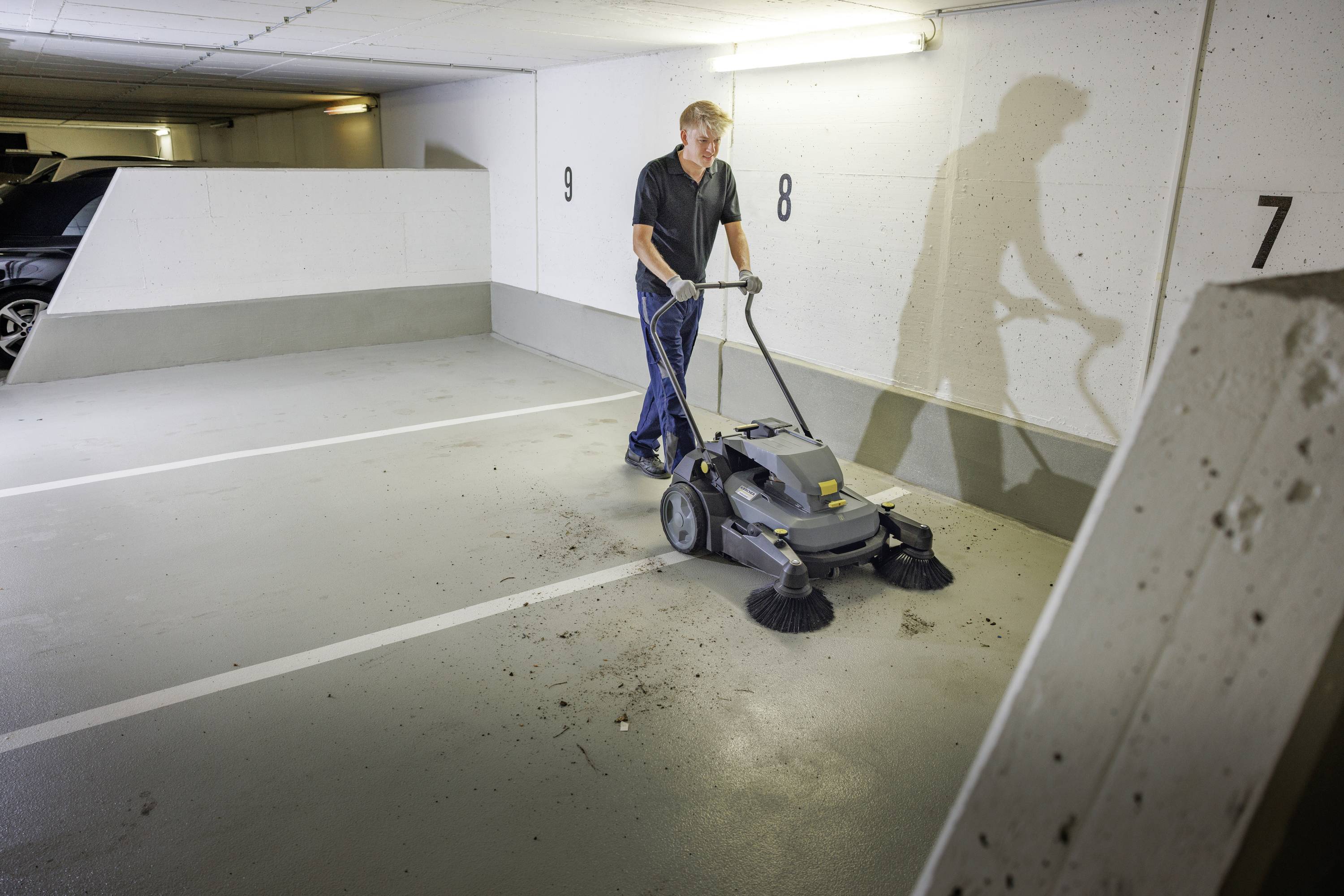 Mann reinigt Parkhausboden mit einer Kehrmaschine, entfernt sichtbaren Schmutz. Weiße Wand mit Parkmarkierungen im Hintergrund.