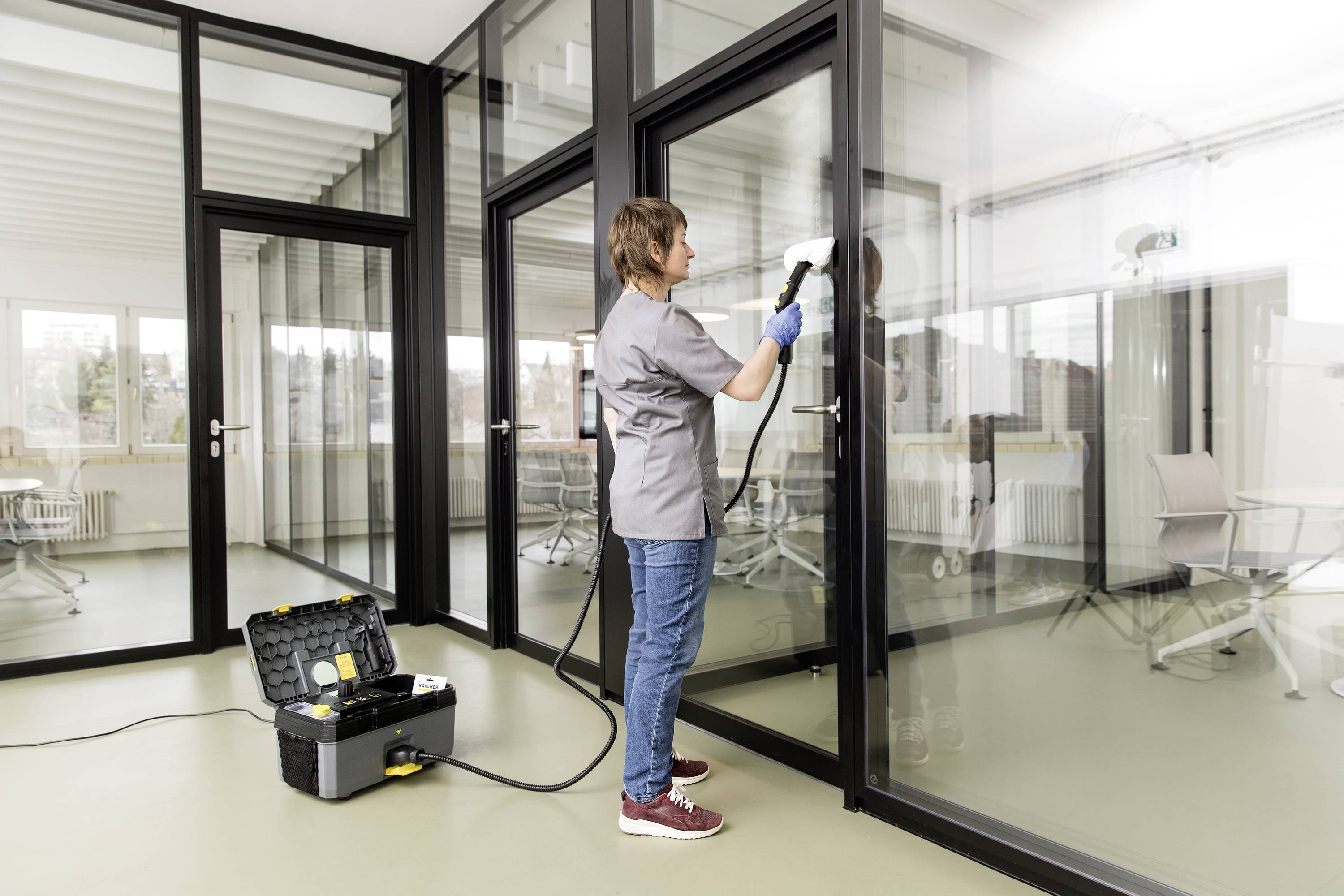Person reinigt mit einem Dampfreiniger eine große Glaswand in einem modernen Büro mit Fenstern im Hintergrund.