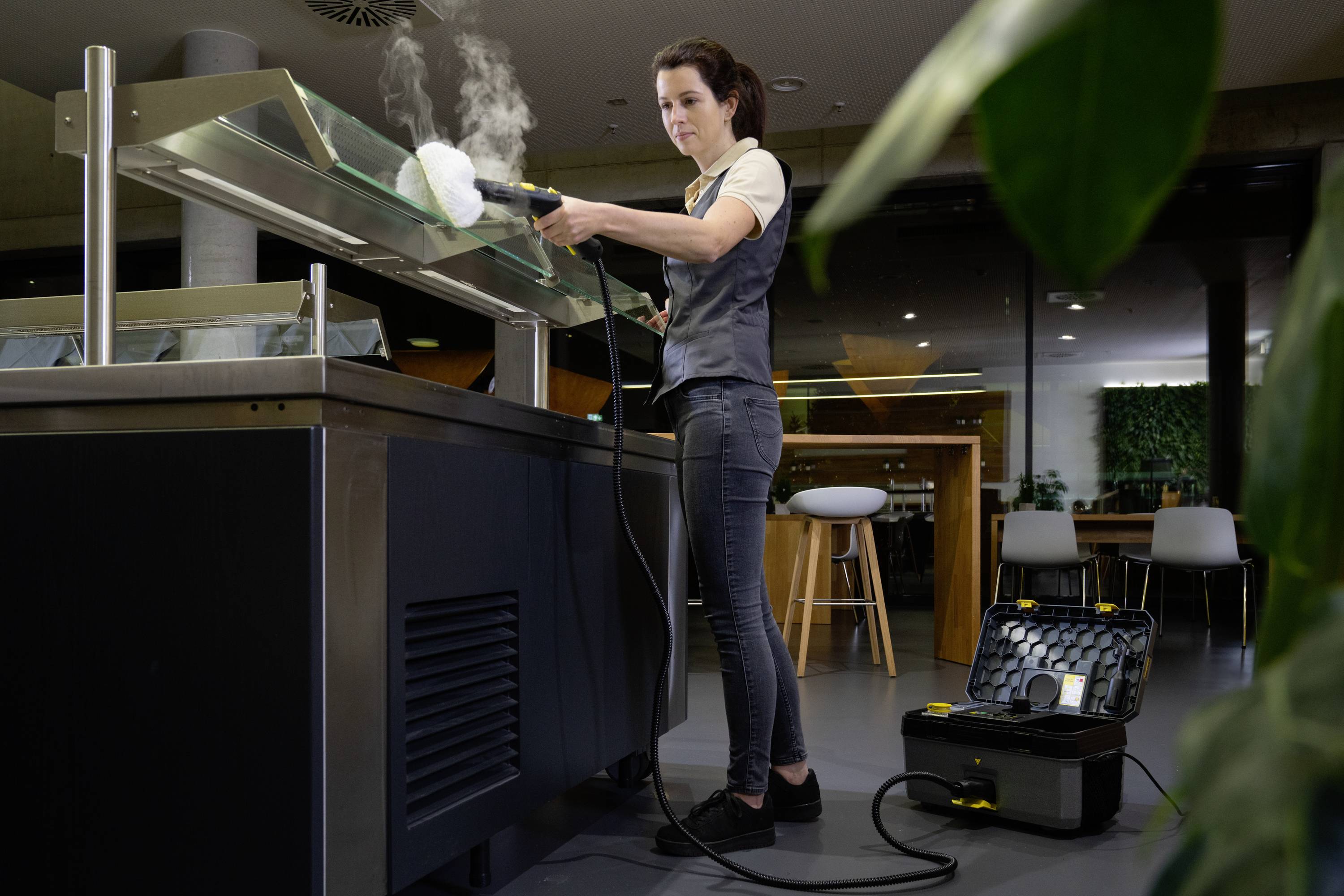 Frau reinigt die Glasscheibe eines Büffets mit einem Dampfreiniger in einem modernen Speisesaal. Reinigungsgerät und Zubehör sind sichtbar.