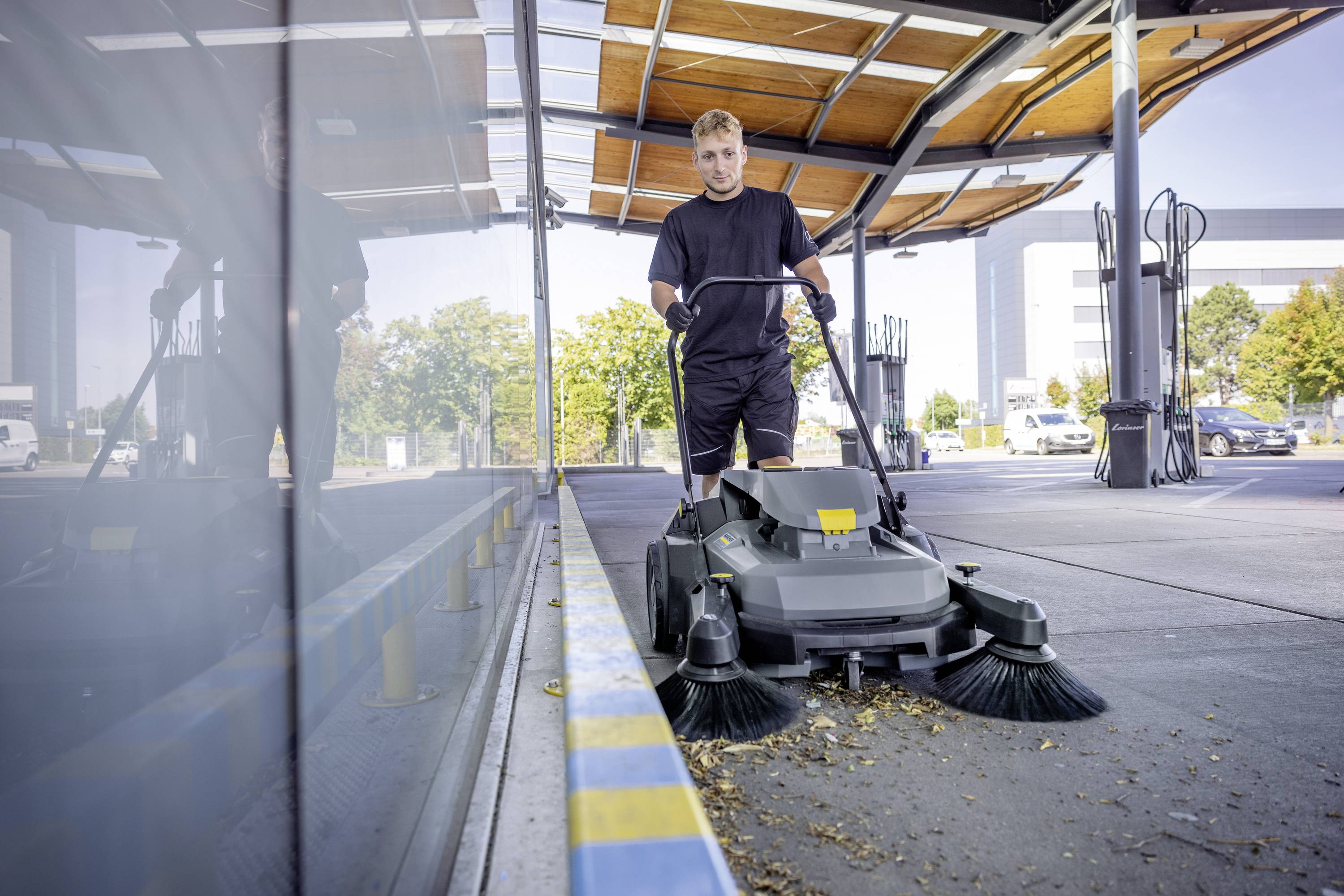 Ein Mann bedient eine elektrische Kehrmaschine in einer offenen Halle. Im Hintergrund sind Bäume und geparkte Autos zu sehen.