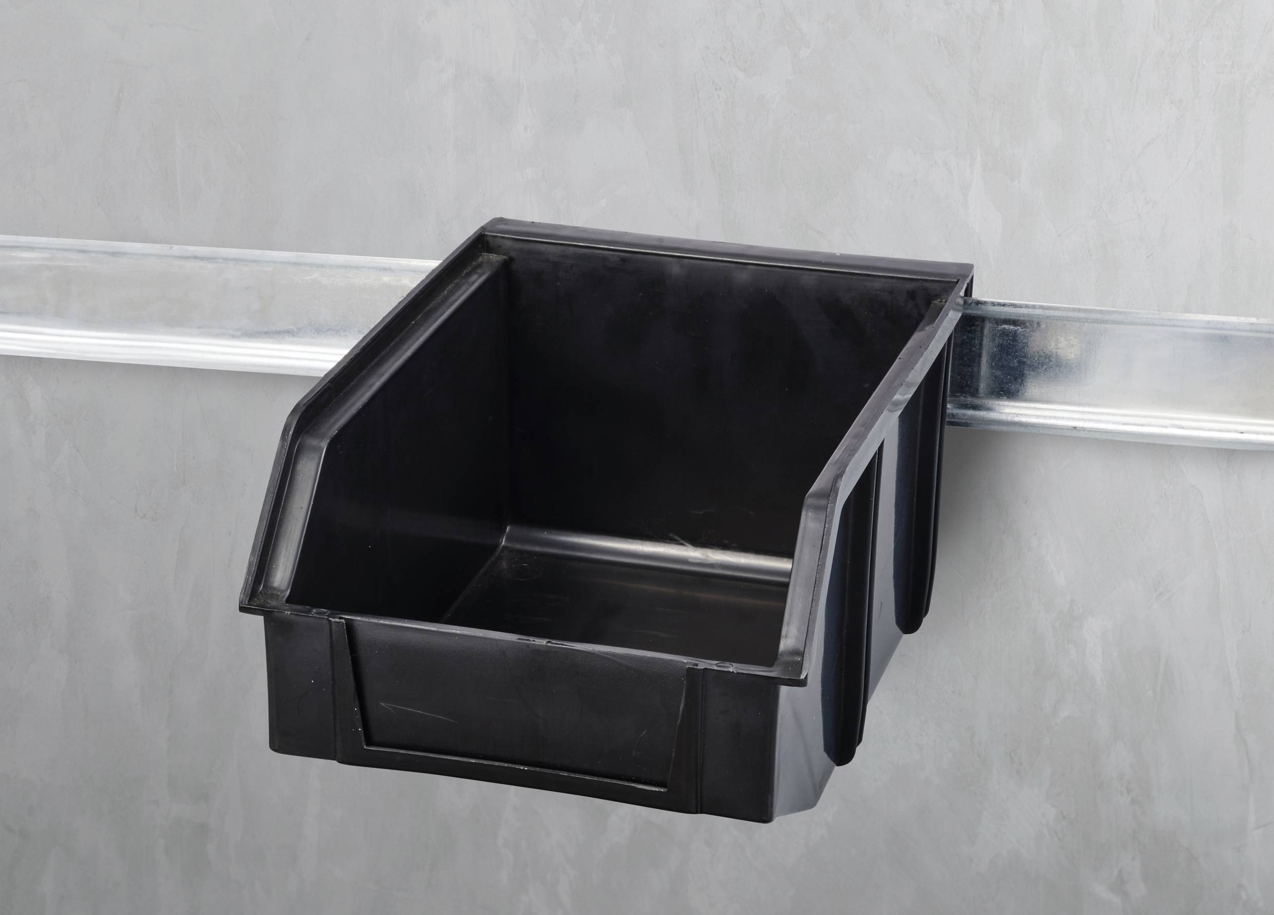 Schwarze Aufbewahrungsbox aus Kunststoff, aufgehängt an einer Metallschiene an der Wand. Box ist leer.
