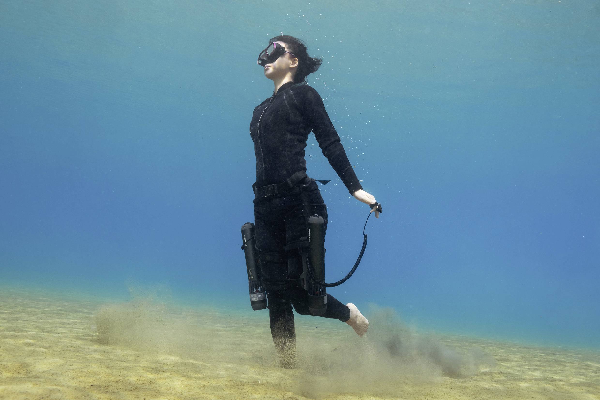 Eine Person in Taucheranzug und Tauchmaske steht unter Wasser auf sandigem Boden und erzeugt Staubwolken. Sie trägt keine Flossen.