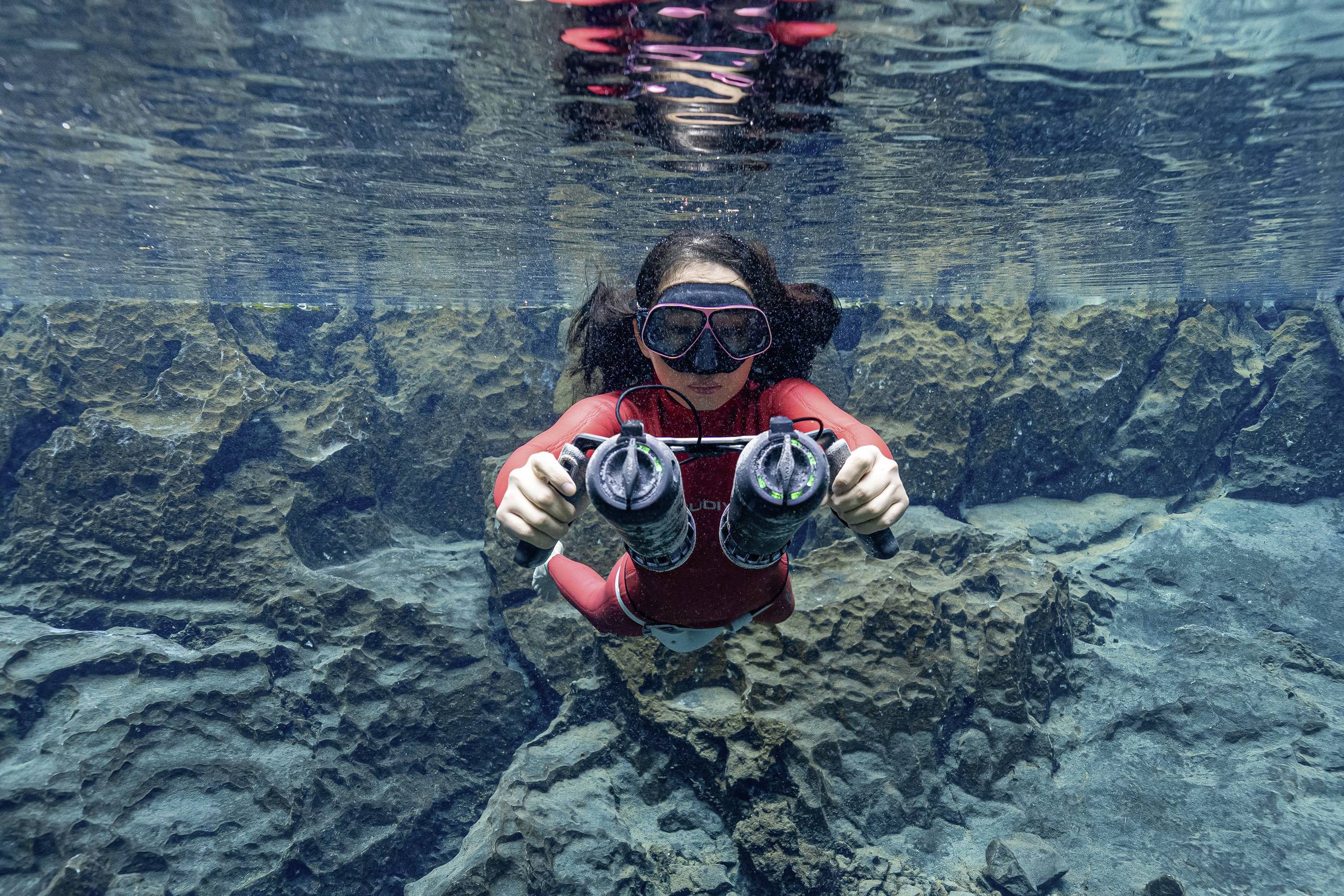 Eine Person taucht unter Wasser in einer klaren Umgebung, hält zwei Geräte, vermutlich Unterwasserkameras, in den Händen.