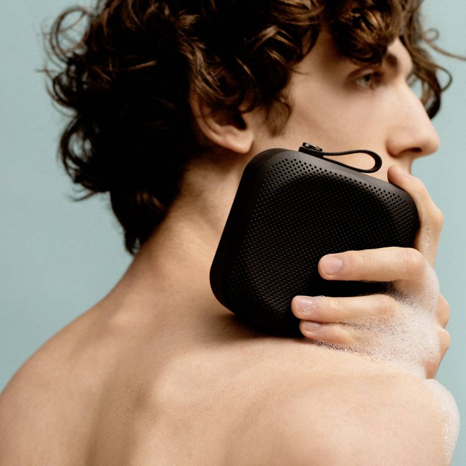 Eine Person hält einen kleinen, schwarzen, tragbaren Lautsprecher an die Schulter. Der Hintergrund ist neutral.