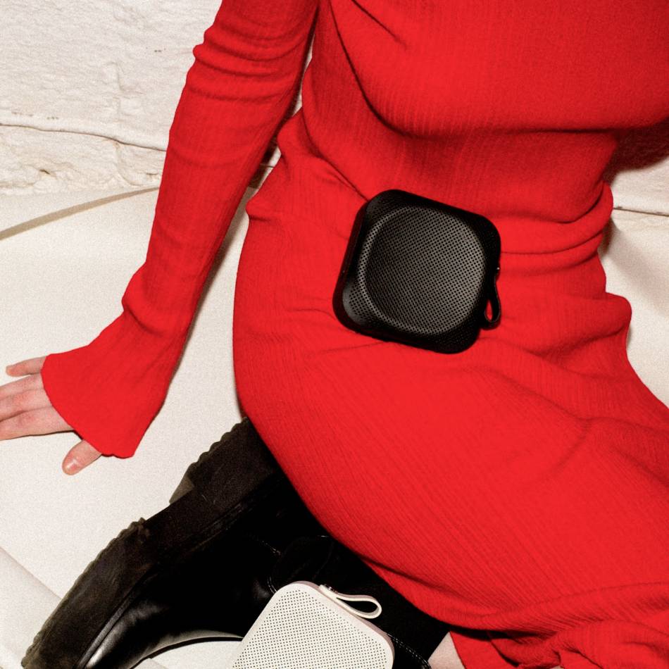 Person in rotem Outfit sitzt auf einem hellen Sofa, hält einen schwarzen und einen weißen tragbaren Lautsprecher.