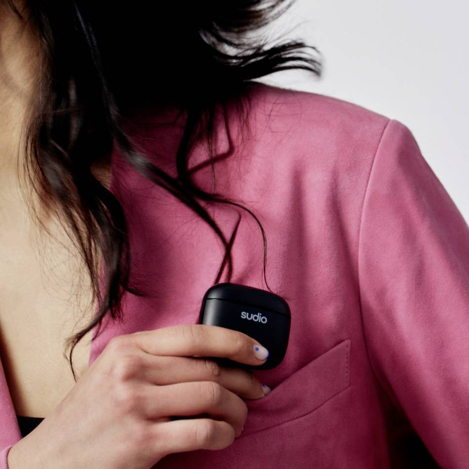 Person mit pinkem Blazer steckt schwarzes Etui mit Logo 'sudio' in die Brusttasche. Haare sind sichtbar, Gesicht nicht.