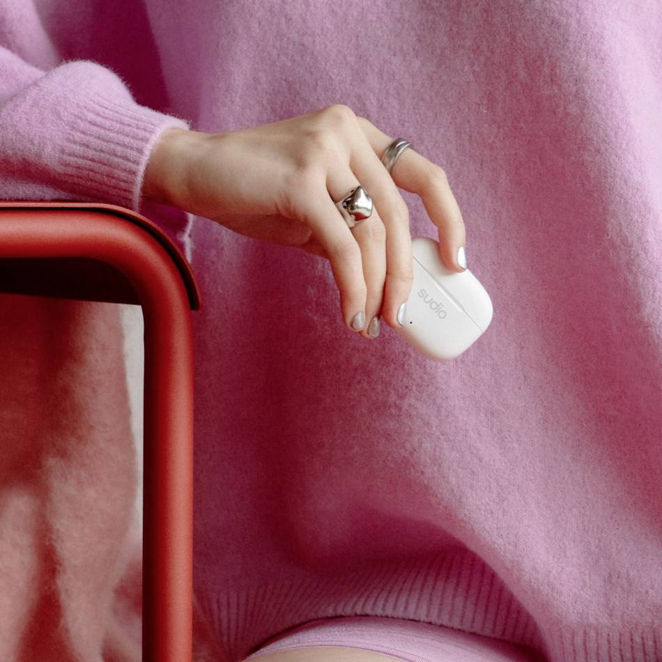 Eine Hand hält ein weißes, kabelloses Ohrhörer-Etui, getragen von einer Person in rosafarbener Kleidung, neben einer roten Stuhllehne.