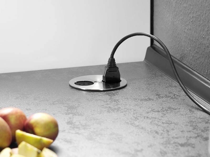 Ein Netzkabel, das in eine runde Steckdose auf einer Küchenarbeitsplatte gesteckt ist, mit Äpfeln und Apfelscheiben in der Nähe, was eine häusliche Umgebung andeutet.