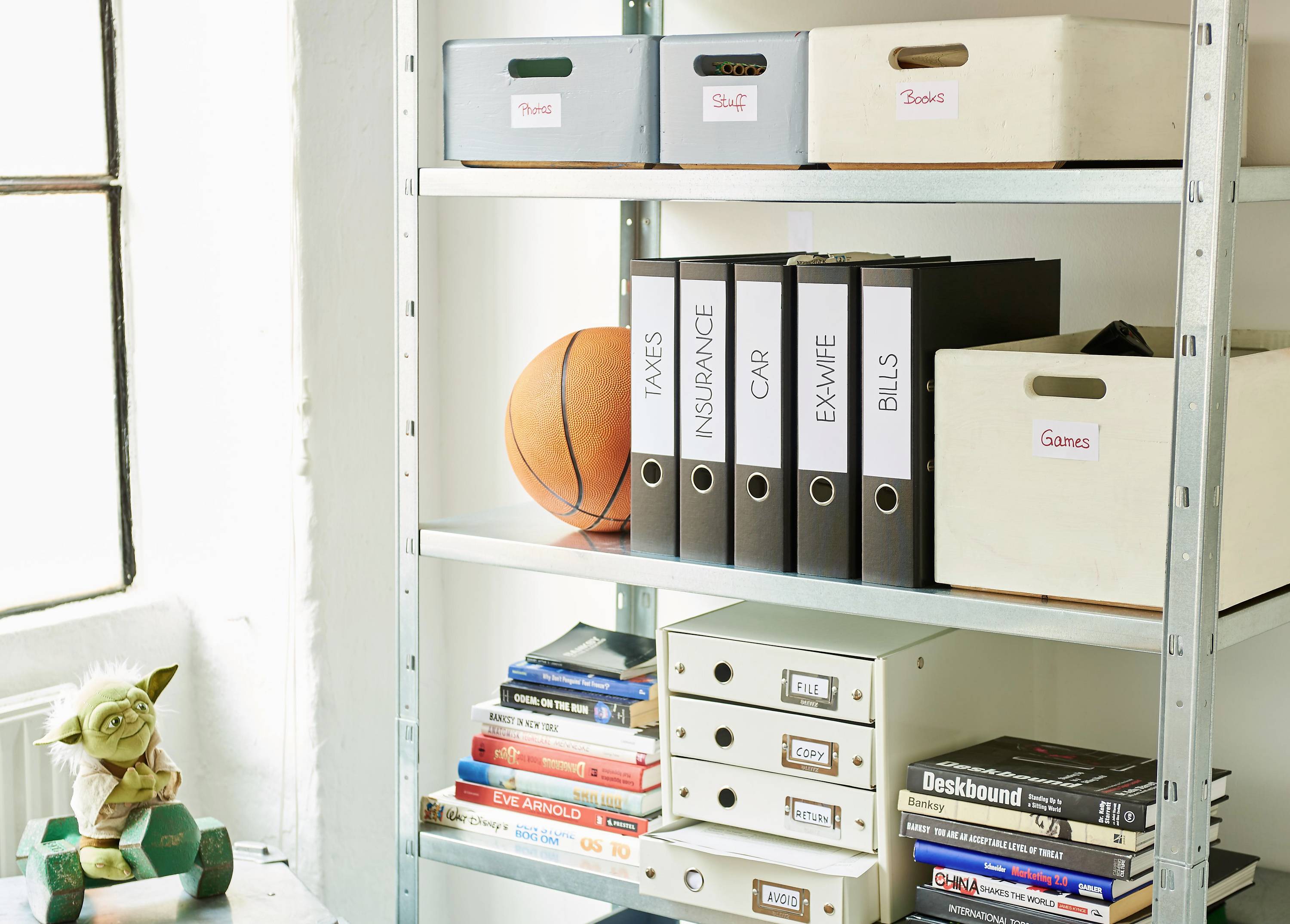 Ein Regal mit Aktenordnern, beschriftet mit 'Tax', 'Insurance', 'Car', daneben Bücherstapel und Spielkartons. Ein Basketball und eine Figur unten.