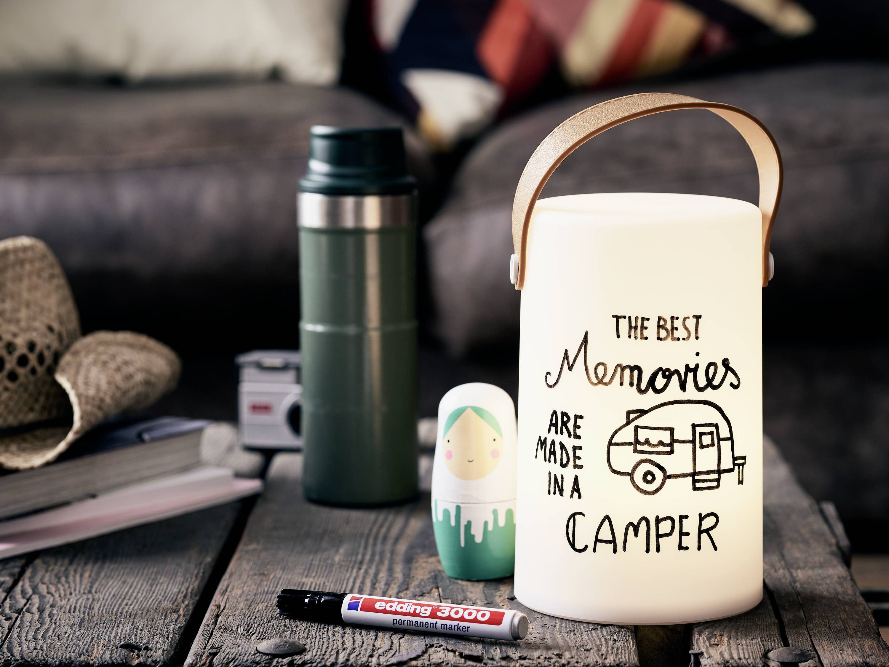 Eine Laterne steht auf einem Tisch mit dem Text 'The best memories are made in a camper'. Im Hintergrund sind weitere Campingutensilien.
