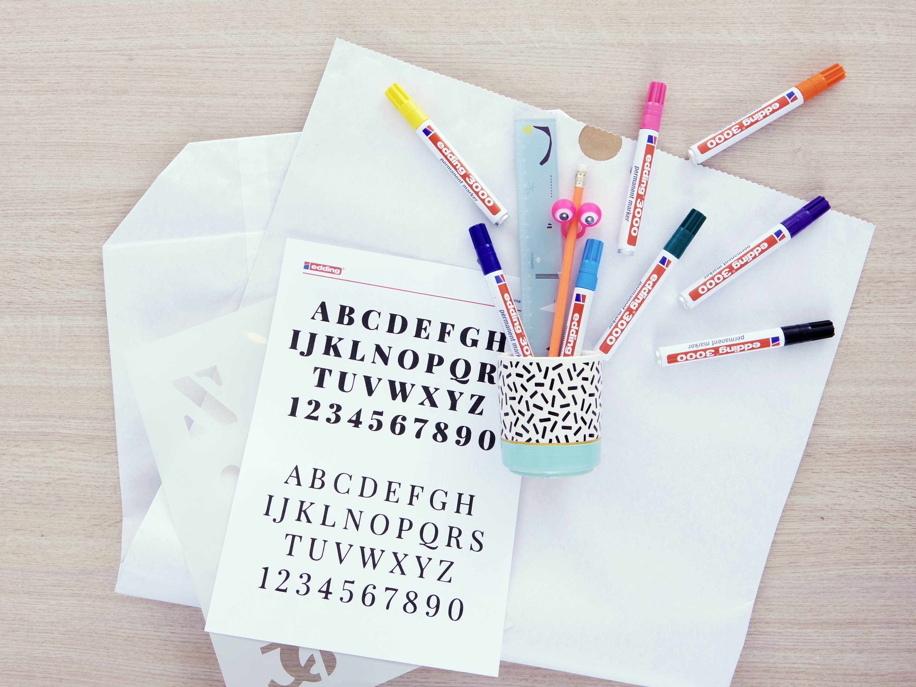 Papier mit Buchstaben und Zahlen liegt neben bunten Stiften in einem Halter. Hintergrund: Holzoberfläche und Umschläge.