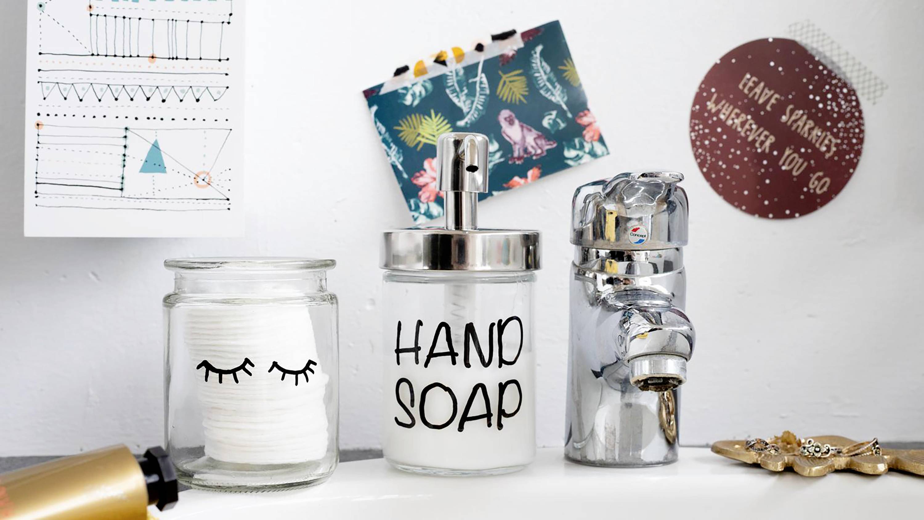 Gläser mit Wattepads, Flüssigseife in einem Spender mit der Aufschrift 'Hand Soap' und Haargummis auf einem Badezimmerregal.