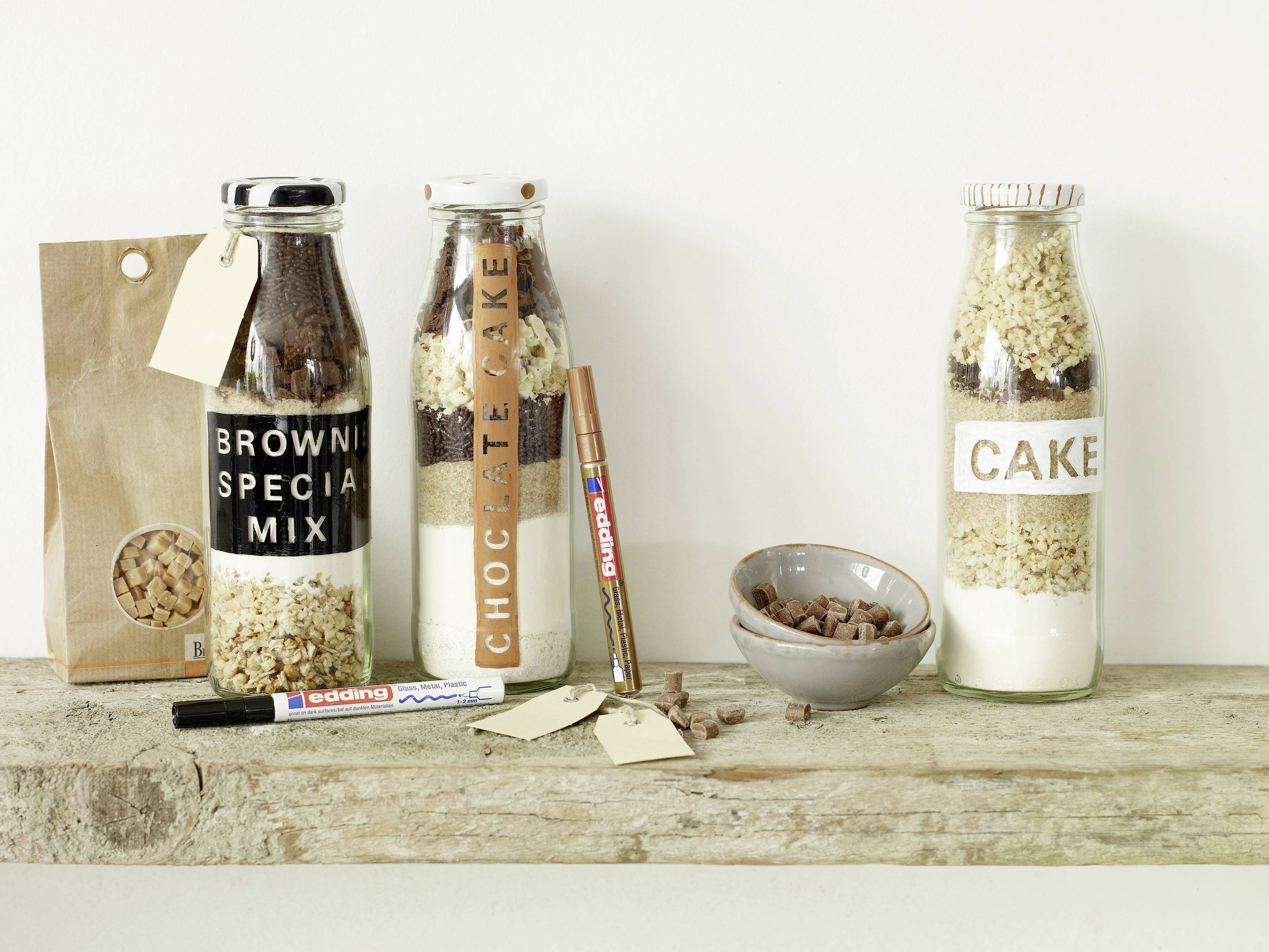 Drei verschiedene Kuchen-Backmischungen in Flaschen auf einem Holzbrett, daneben eine Schüssel mit Kakaobohnen und verschiedene Beschriftungsmaterialien.