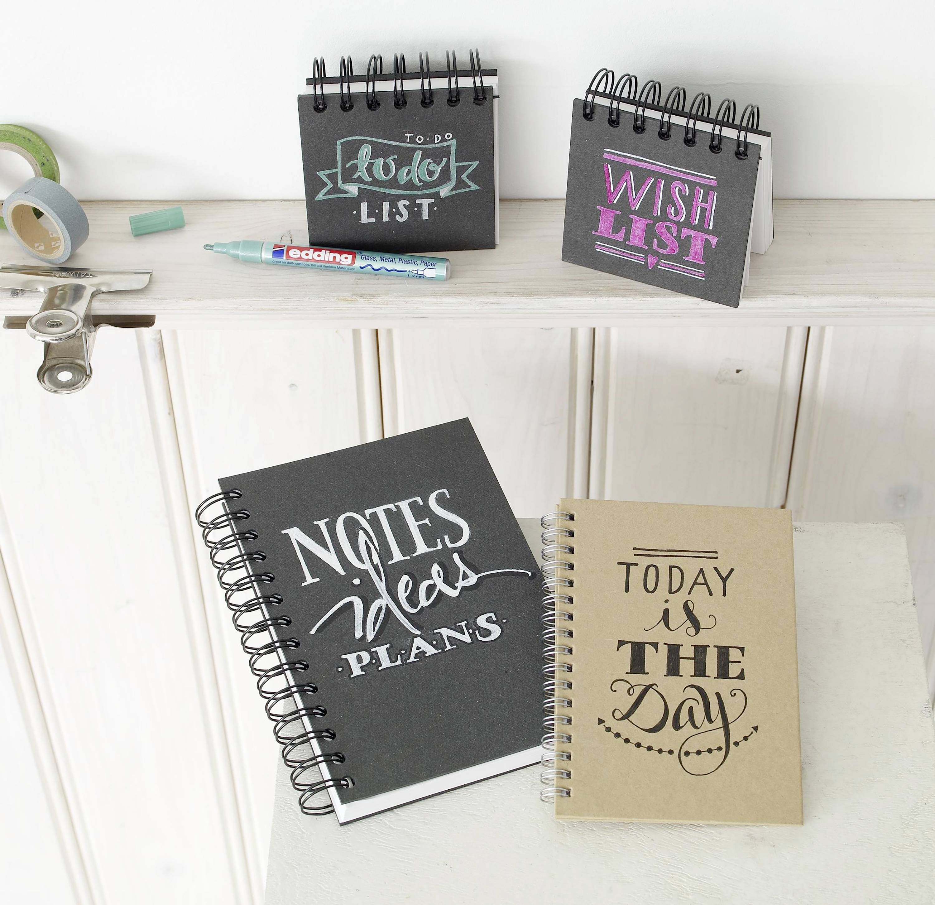 Fünf Notizbücher auf einem Holzuntergrund. Die Titel lauten: 'To Do List', 'Wish List', 'Plans - Notes - Ideas' und 'Today is the Day'. Schreibutensilien sind daneben platziert.