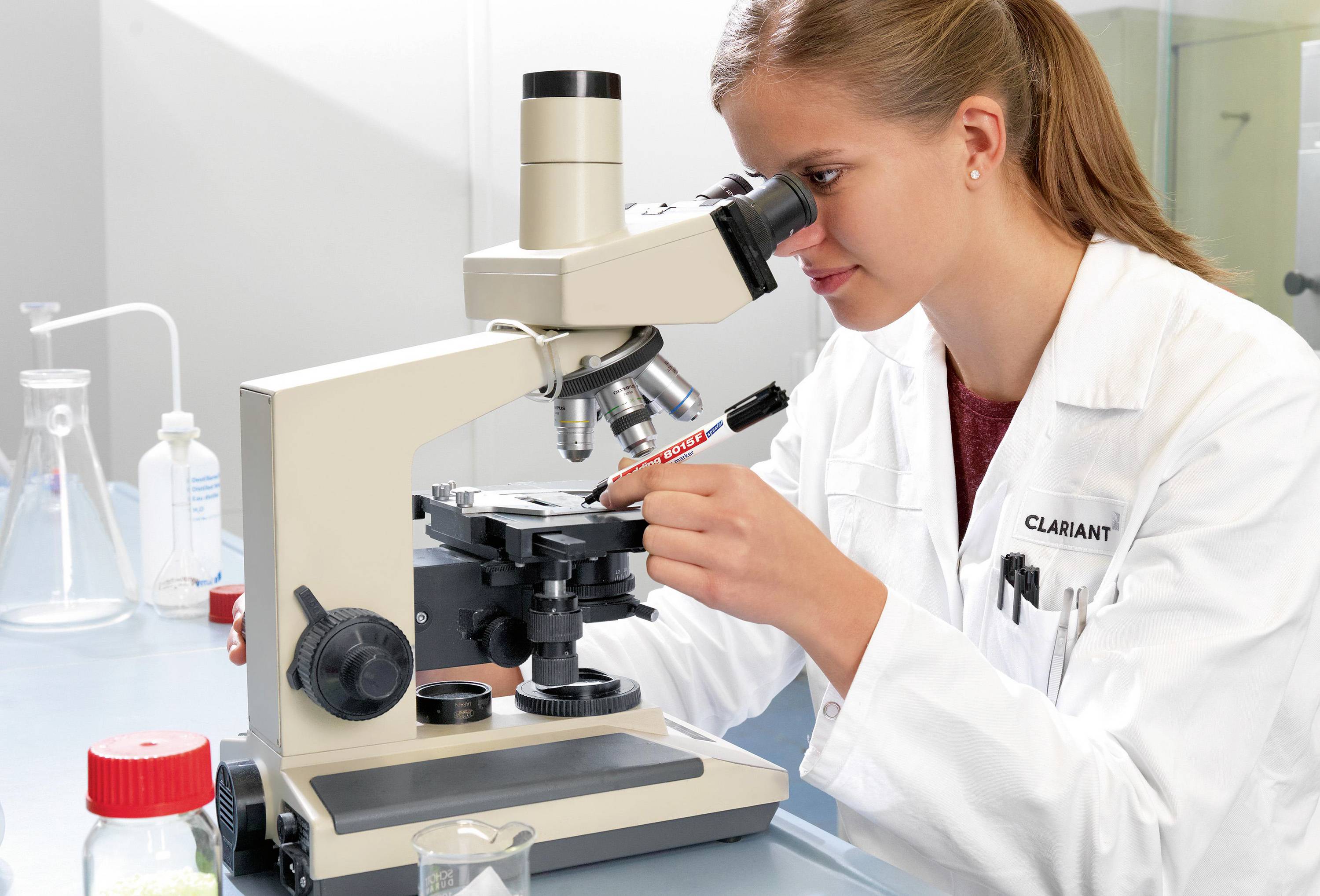 Eine Frau im weißen Kittel benutzt ein Mikroskop in einem Labor. Im Hintergrund sind Glasflaschen und Laborausrüstung zu sehen.