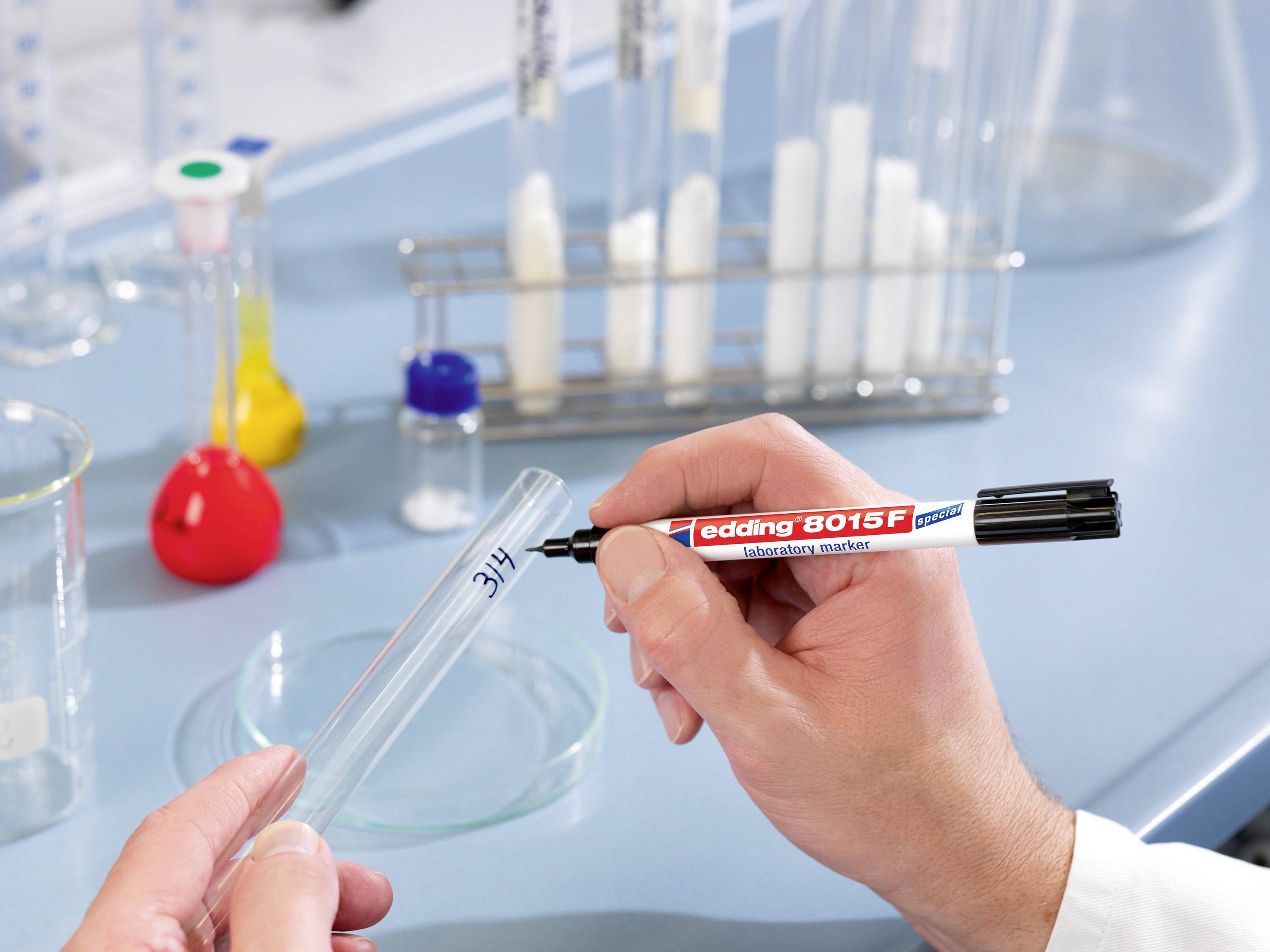Eine Hand beschriftet ein Reagenzglas mit einem schwarzen Marker im Labor. Im Hintergrund sind weitere Labormaterialien sichtbar.